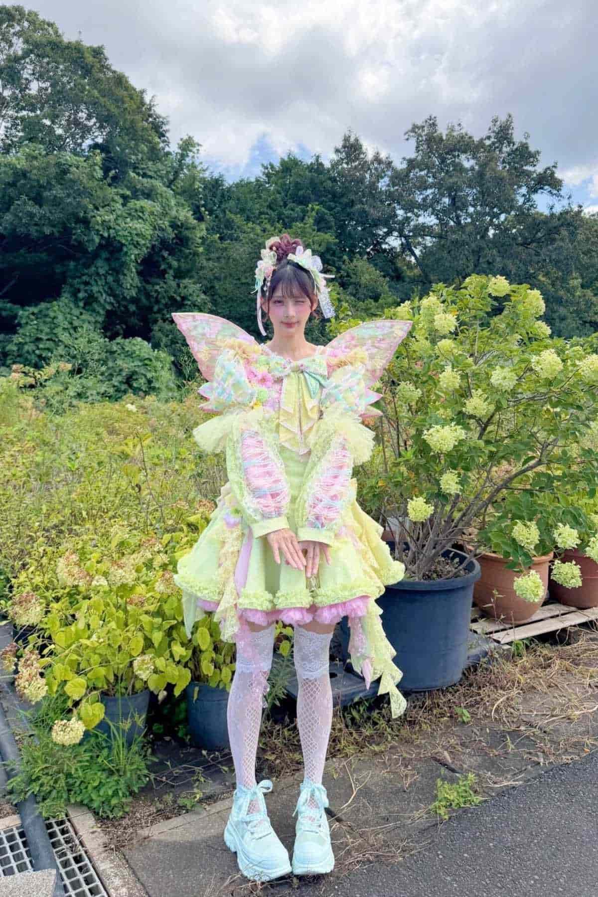Person standing outdoors wearing a pastel fairy-inspired costume with wings, lace stockings, and platform shoes, surrounded by green plants and shrubs.