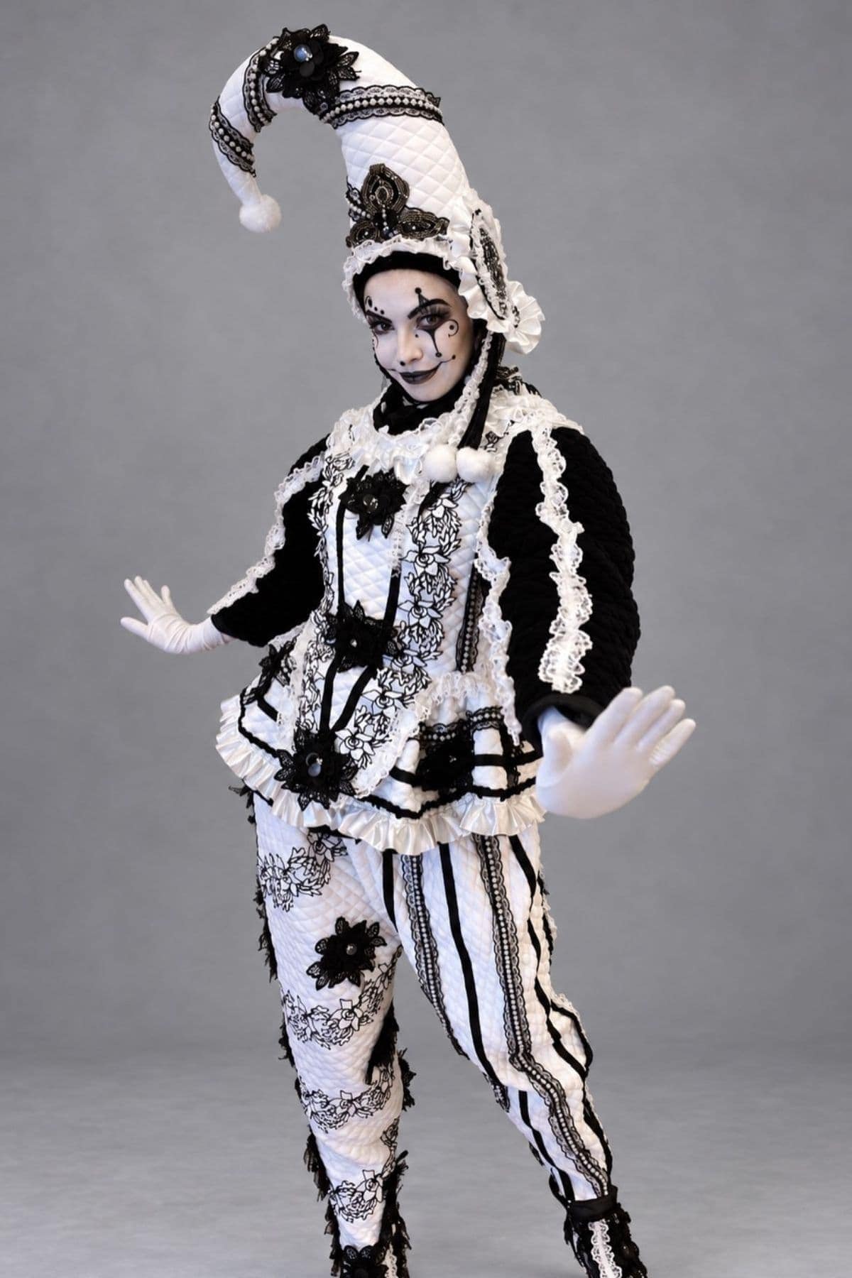 Person in an elaborate black and white harlequin costume with face paint, ruffled details, and a tall, pointed hat posing against a plain gray background.