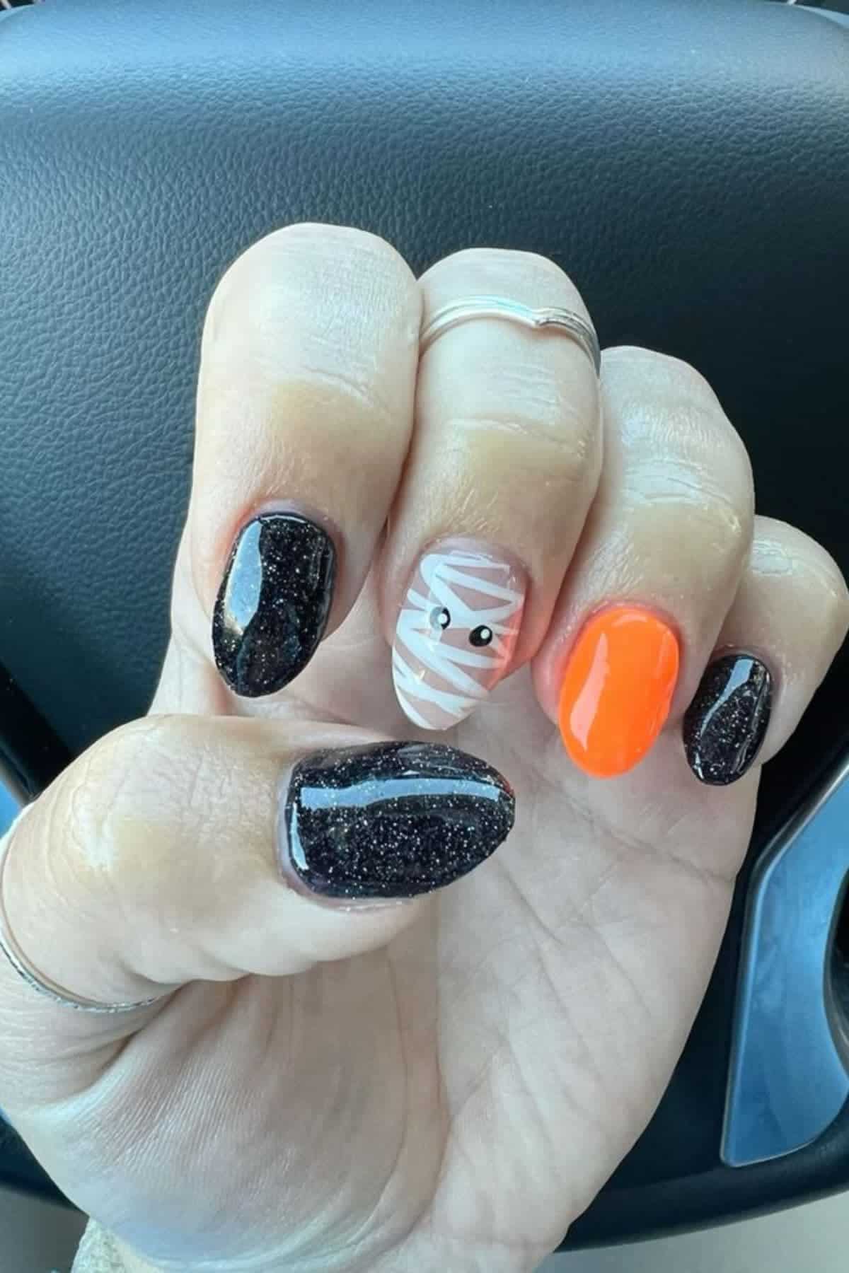 A hand with four black glitter nails, one bright orange nail, and one nail with a white mummy design featuring two small eyes, shown in front of a car steering wheel.