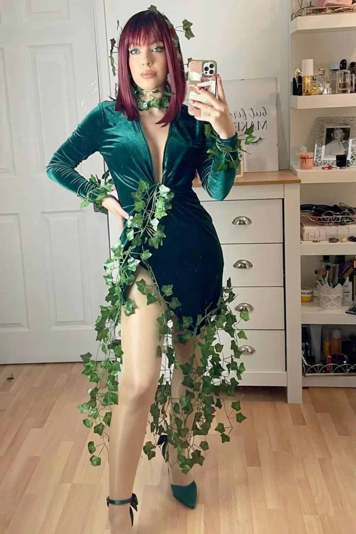 Person in a green dress with artificial ivy draped around them, taking a mirror selfie in a bedroom with wooden floor and white furniture.