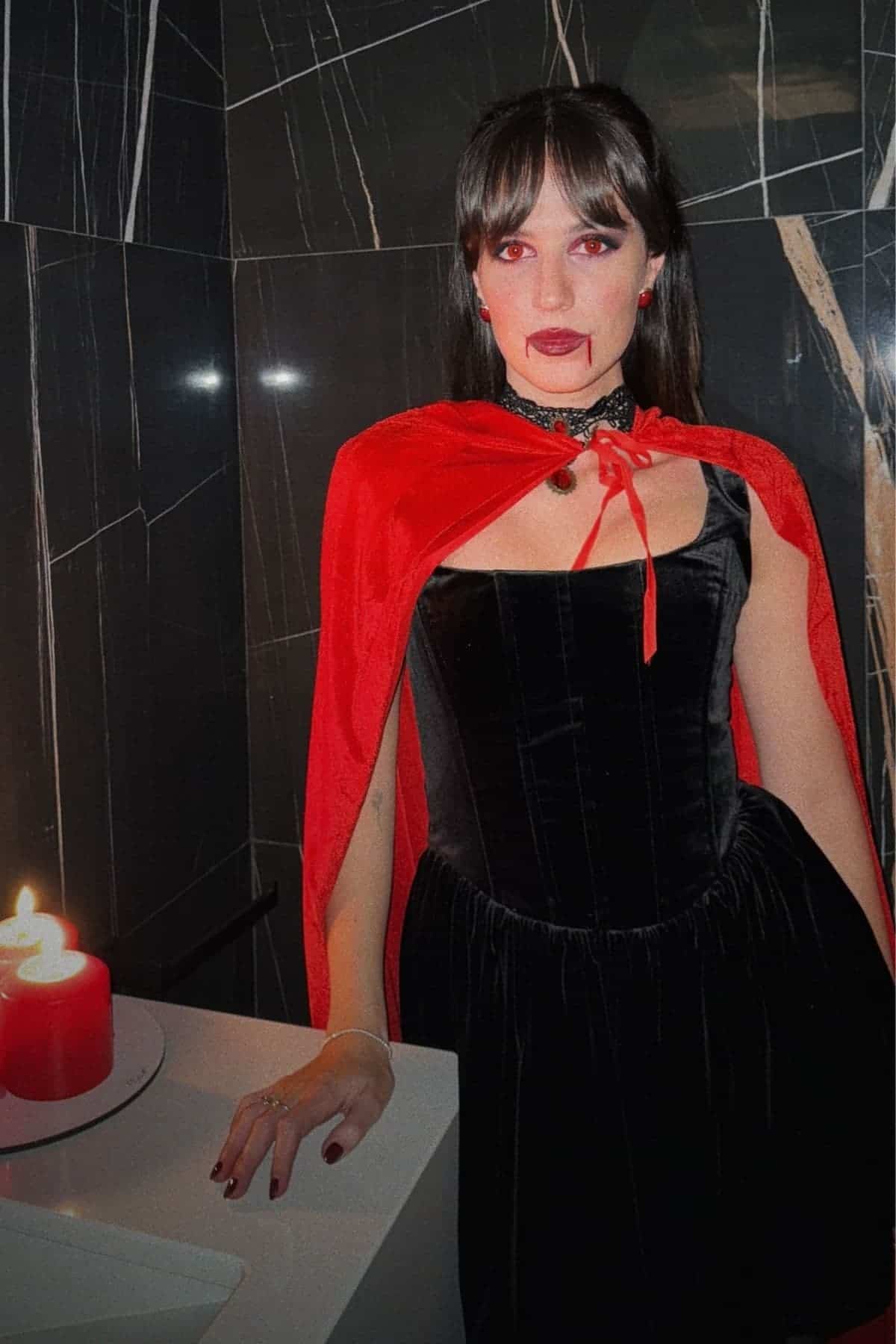 Woman in a black velvet dress and red cape with vampire makeup stands in a dark marble bathroom beside two lit red candles.