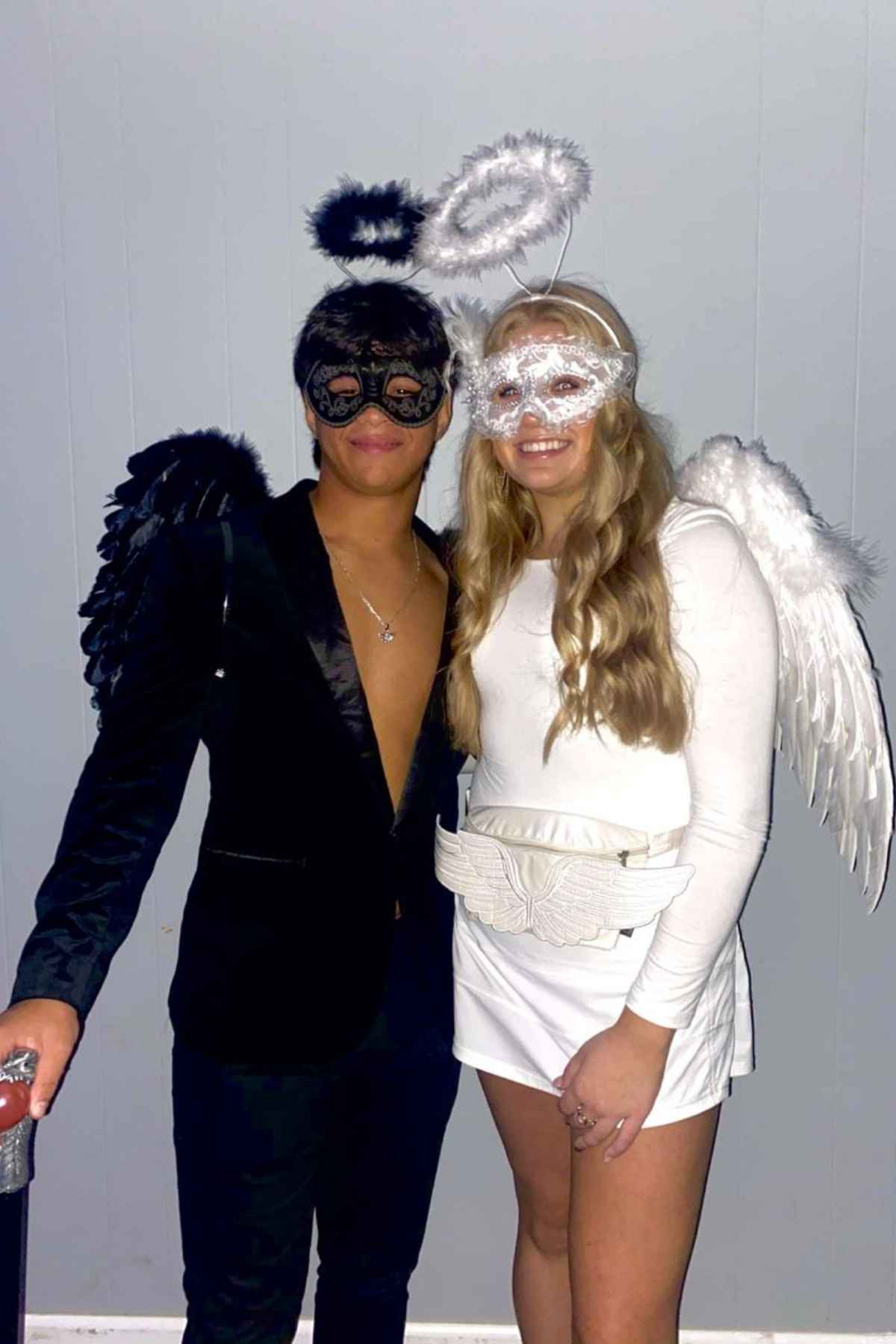 Two people in black and white angel costumes with wings, halos, and masks stand side by side against a plain gray wall.