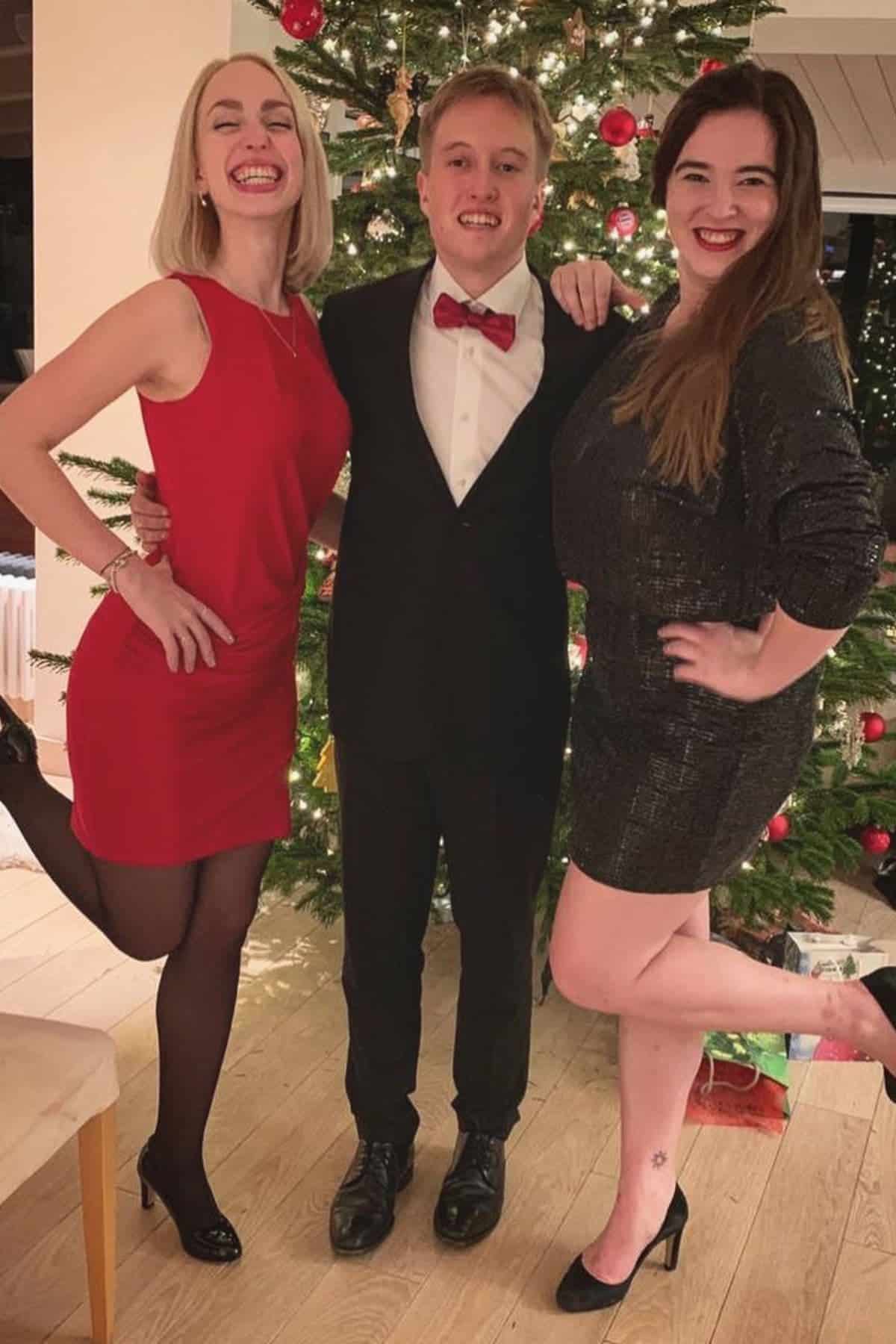 Three people in formal attire pose and smile in front of a decorated Christmas tree; two women wear dresses and heels, and a man stands between them in a suit with a red bow tie.