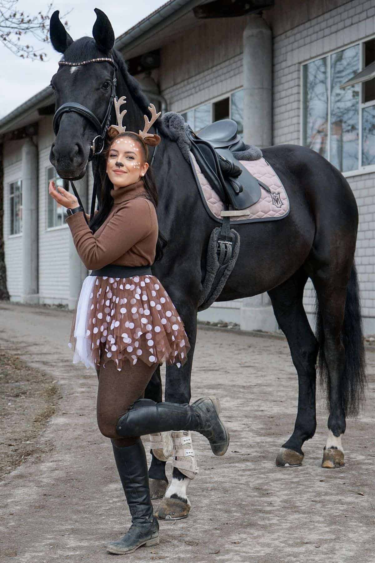 A woman in a brown and white reindeer costume poses with a black horse near a brick building, lifting one foot and smiling at the camera.