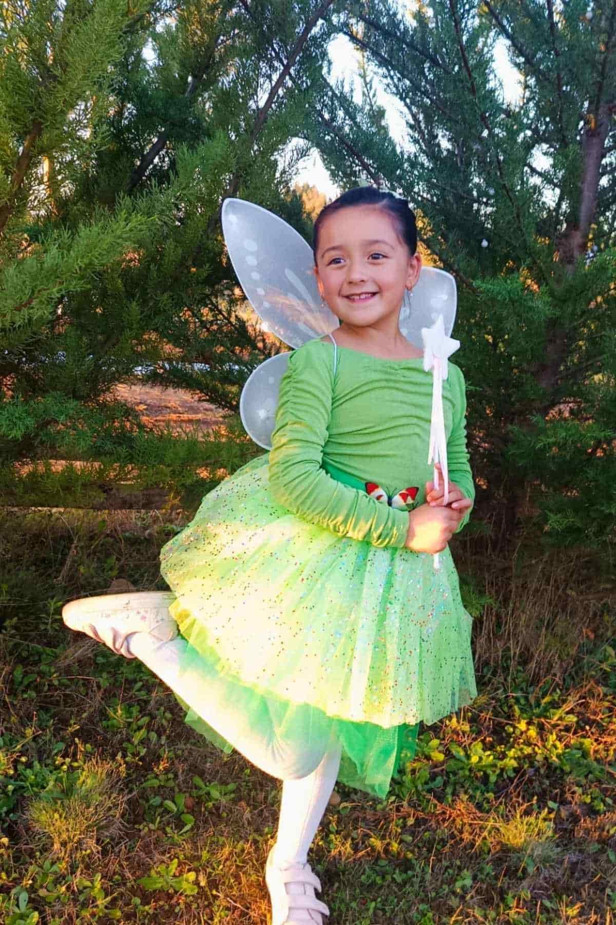 Young child in a green fairy costume with wings and a wand, standing outdoors near green bushes, smiling with one leg raised.