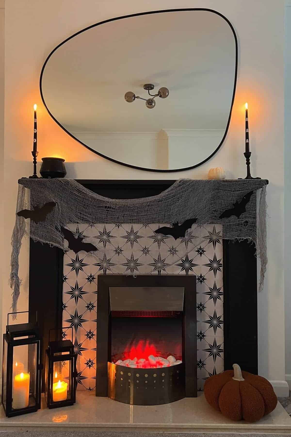 A fireplace decorated with black candles, a gray cloth with bat cutouts, lanterns, a faux fire, and a knitted pumpkin, with a large irregular mirror above.