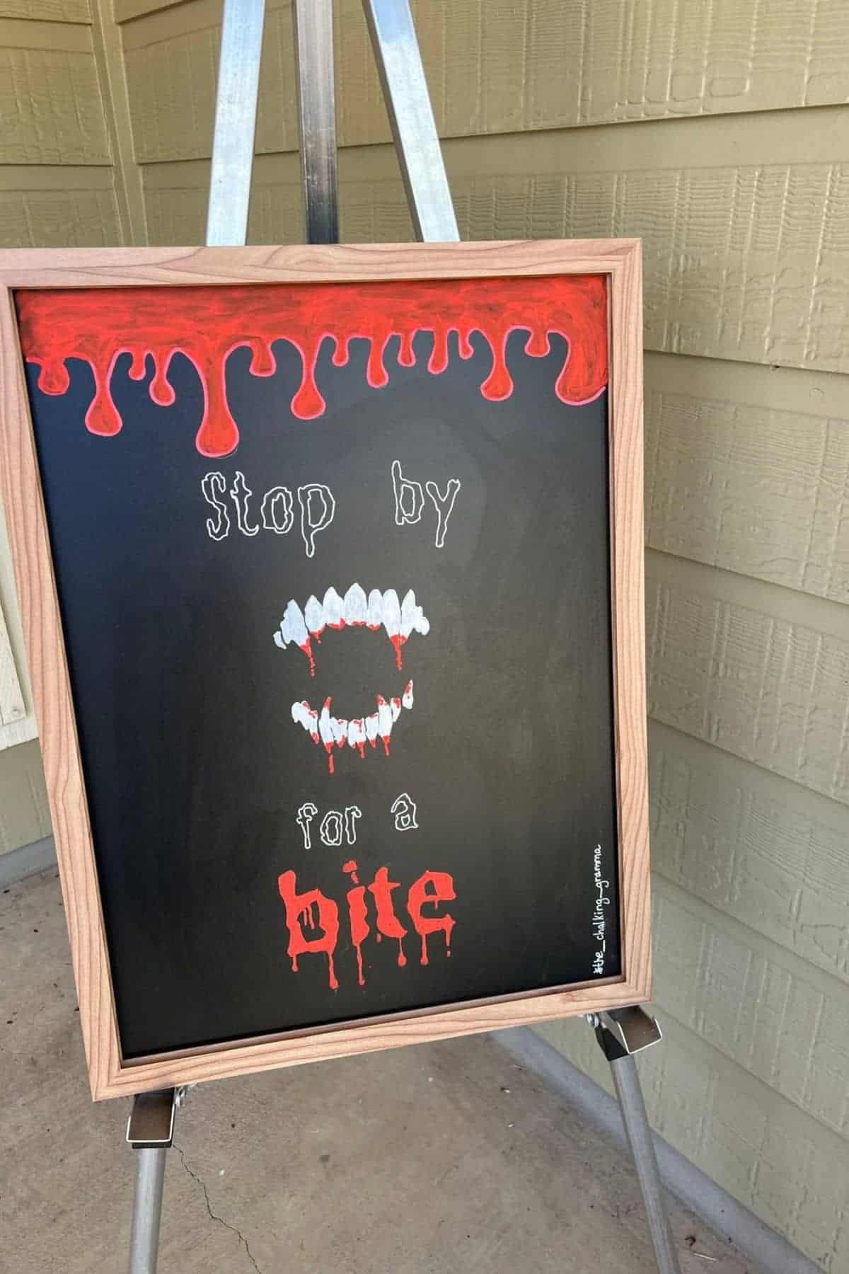 Chalkboard sign with dripping red paint, white vampire fangs, and text: “Stop by for a bite.” Sign is propped on an easel against a wall.