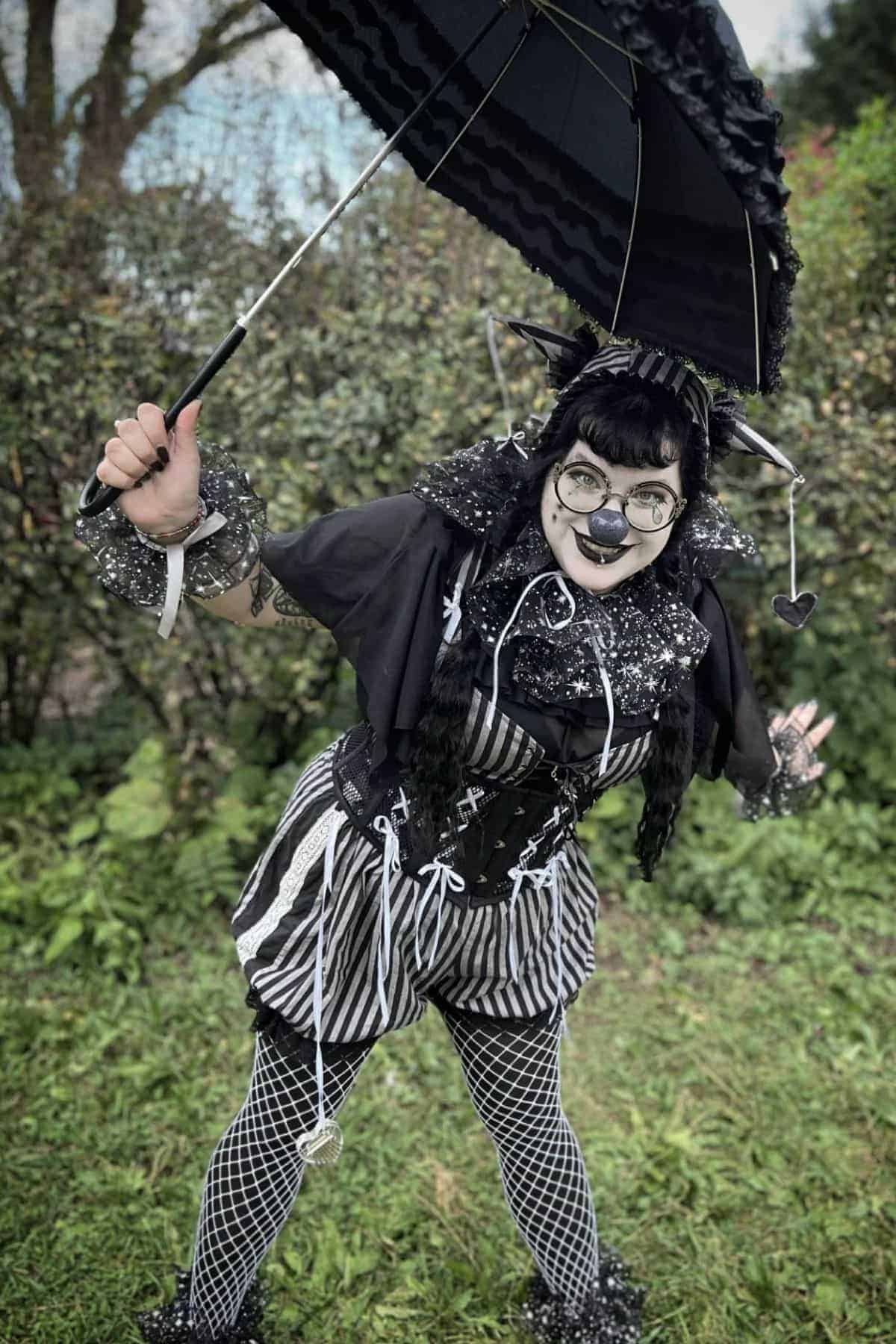 Person in black and white striped costume with face paint holds a black umbrella, standing outdoors on grass with bushes in the background.