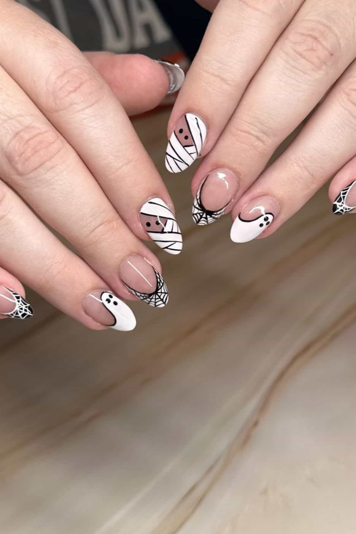 Hands with almond-shaped nails featuring Halloween-themed nail art, including ghost, mummy, and spiderweb designs in white, black, and nude tones.