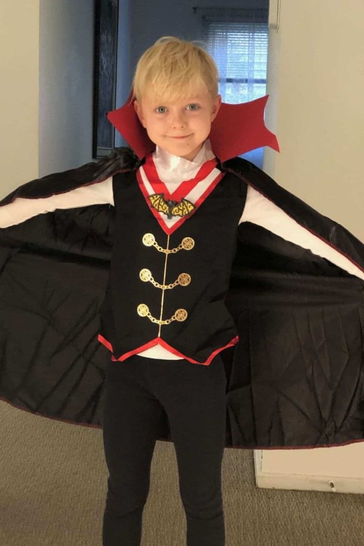 Child wearing a vampire costume with a black cape, red collar, white shirt, vest, and medallion, standing indoors with arms spread open.