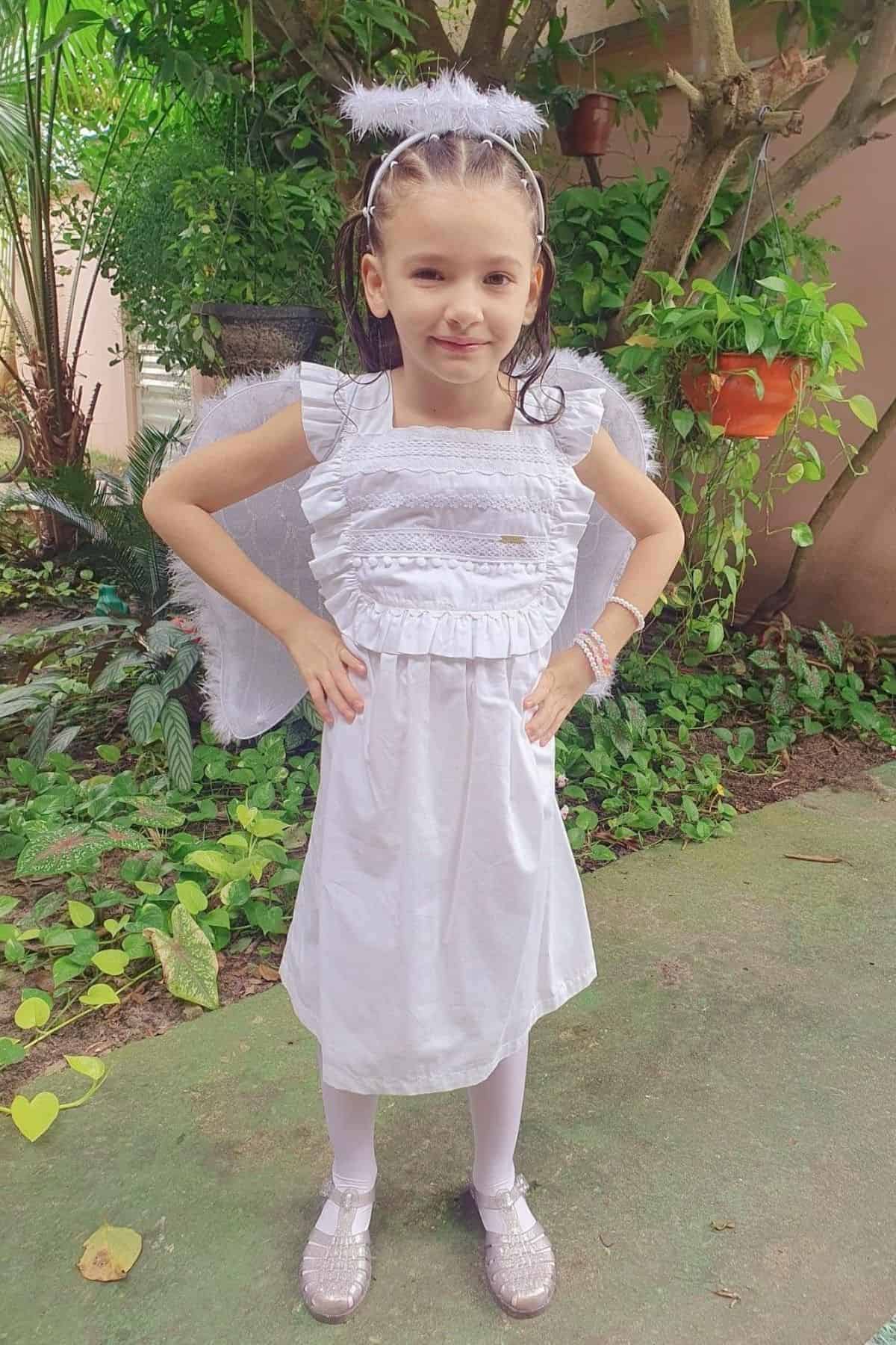 Young girl dressed as an angel in a white dress, wings, and halo stands outdoors on a garden path with hands on her hips.
