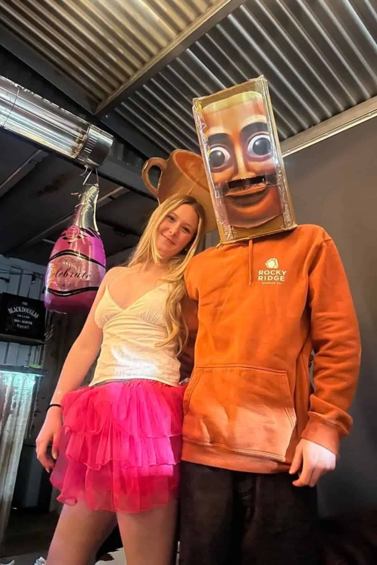 Two people pose indoors; one wears a white top with a pink tutu, the other wears an orange hoodie and a box with painted eyes and mouth as a mask. Inflatable bottle props hang nearby.