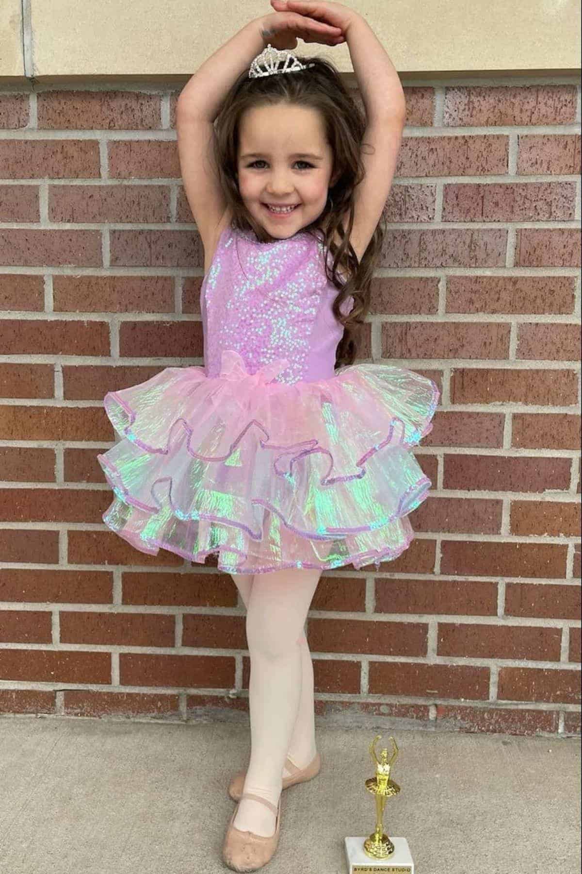 Young child in a sparkly pink ballet costume and tiara poses on one foot in front of a brick wall, smiling, with a trophy on the ground by her feet.
