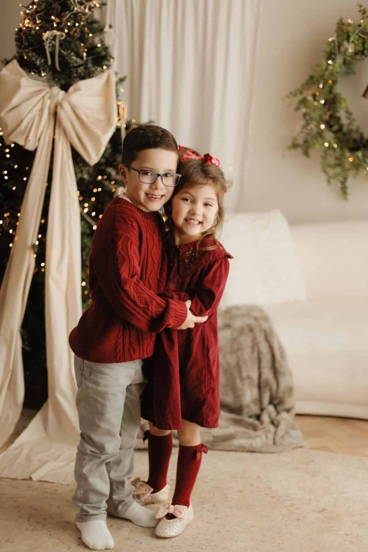 Two young children in festive red outfits stand hugging and smiling in front of a decorated Christmas tree and holiday decor.