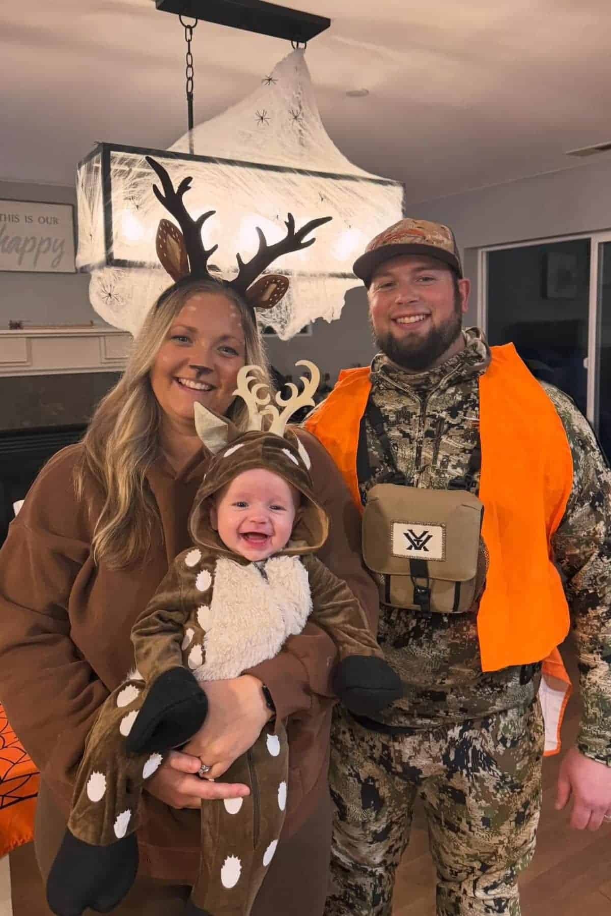 Three people pose indoors: an adult woman and a baby dressed as deer, and an adult man dressed in camouflage hunting gear with an orange vest.