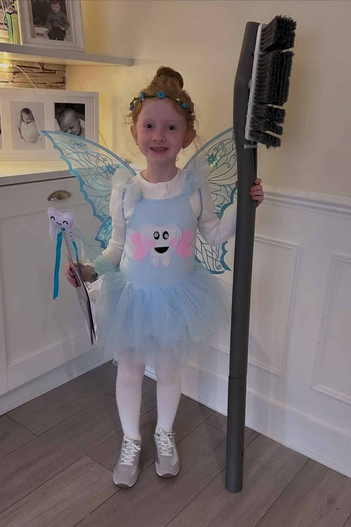 Young child in a tooth fairy costume with wings, holding a large toothbrush prop in one hand and a tooth wand in the other, standing indoors and smiling.