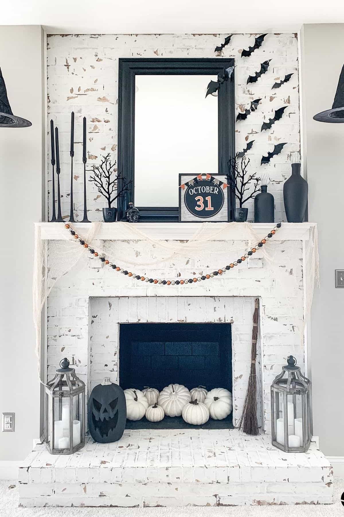 White brick fireplace decorated for Halloween with black bats, candles, a “October 31” sign, white pumpkins, a broom, lanterns with candles, and black hanging hats.