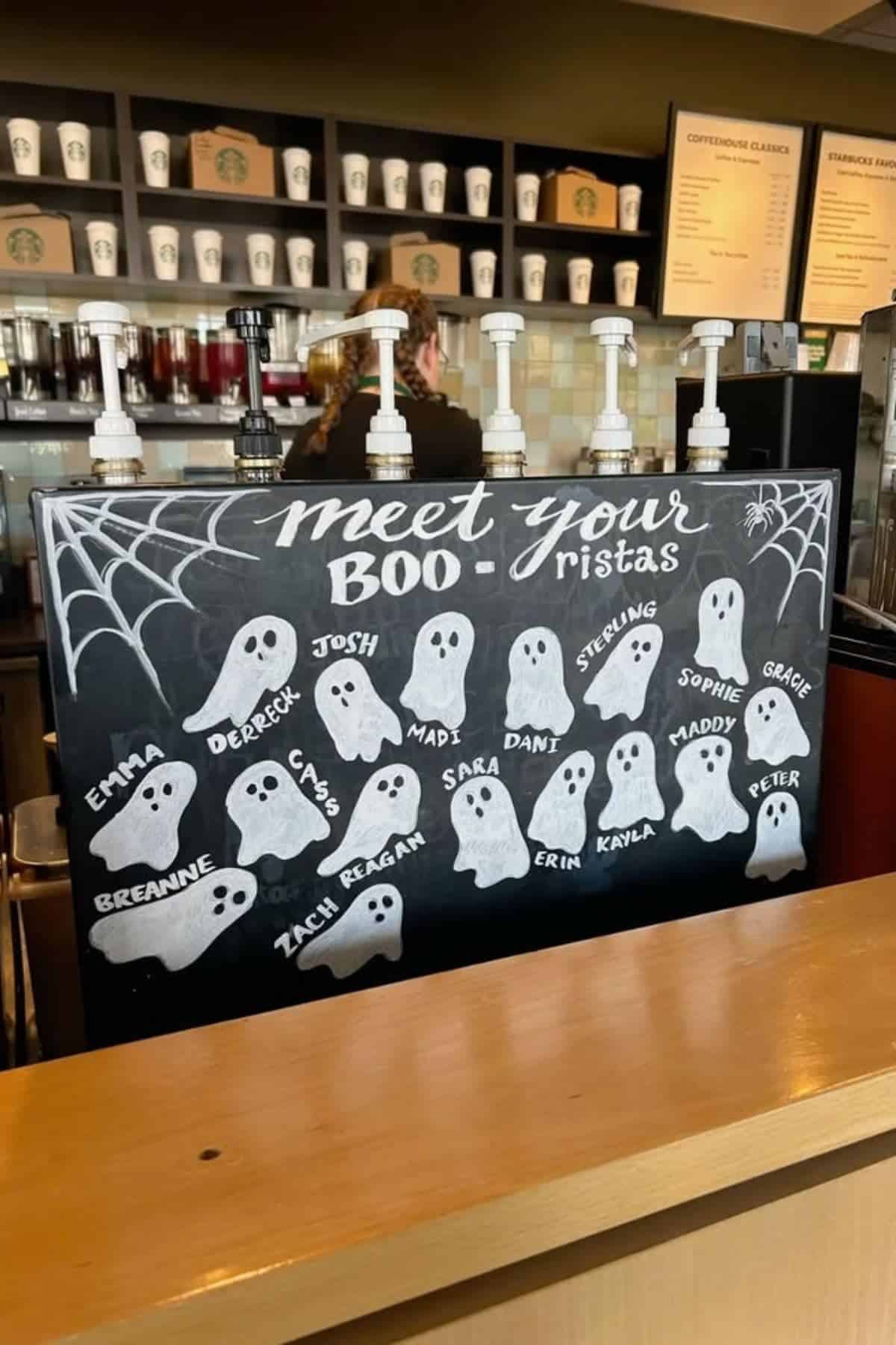 A chalkboard at a coffee shop displays ghost drawings with staff names under the heading “Meet Your Boo-ristas.” Syrup pumps and cups are visible in the background.