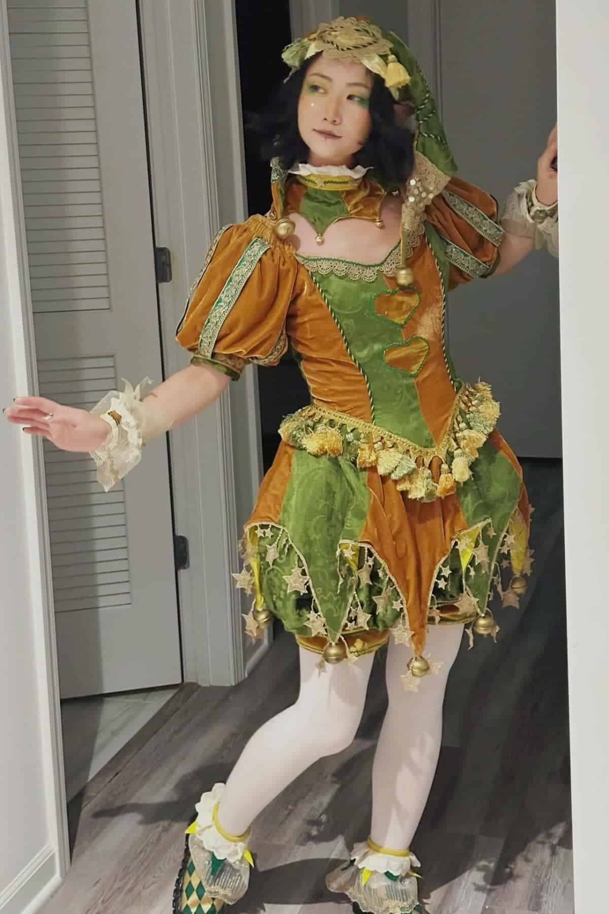 Person in an elaborate green and orange costume with puffed sleeves and a pointed hat, standing indoors on a wood floor near a doorway.
