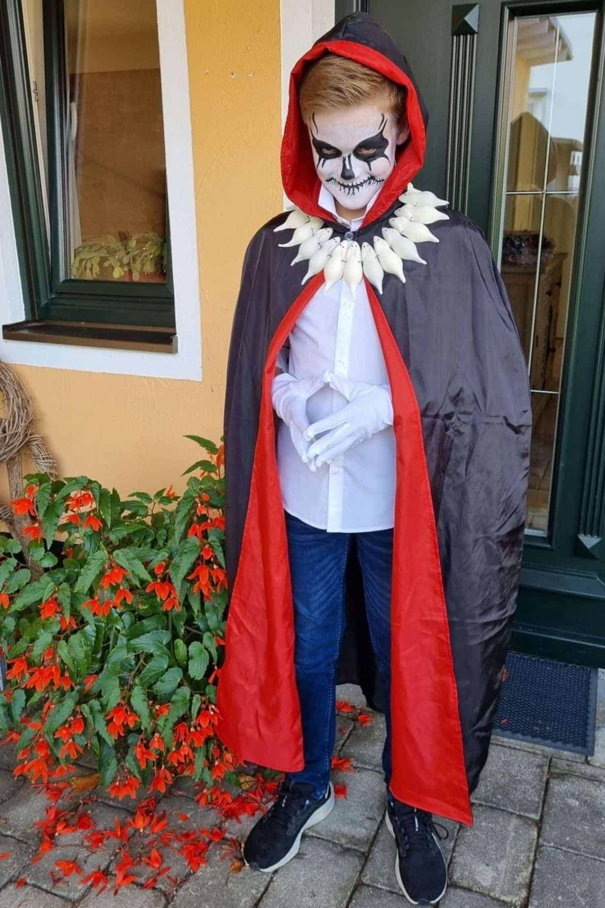 Person wearing a black and red cape, skull makeup, white gloves, and a bone necklace stands outside near a plant and a house entrance.