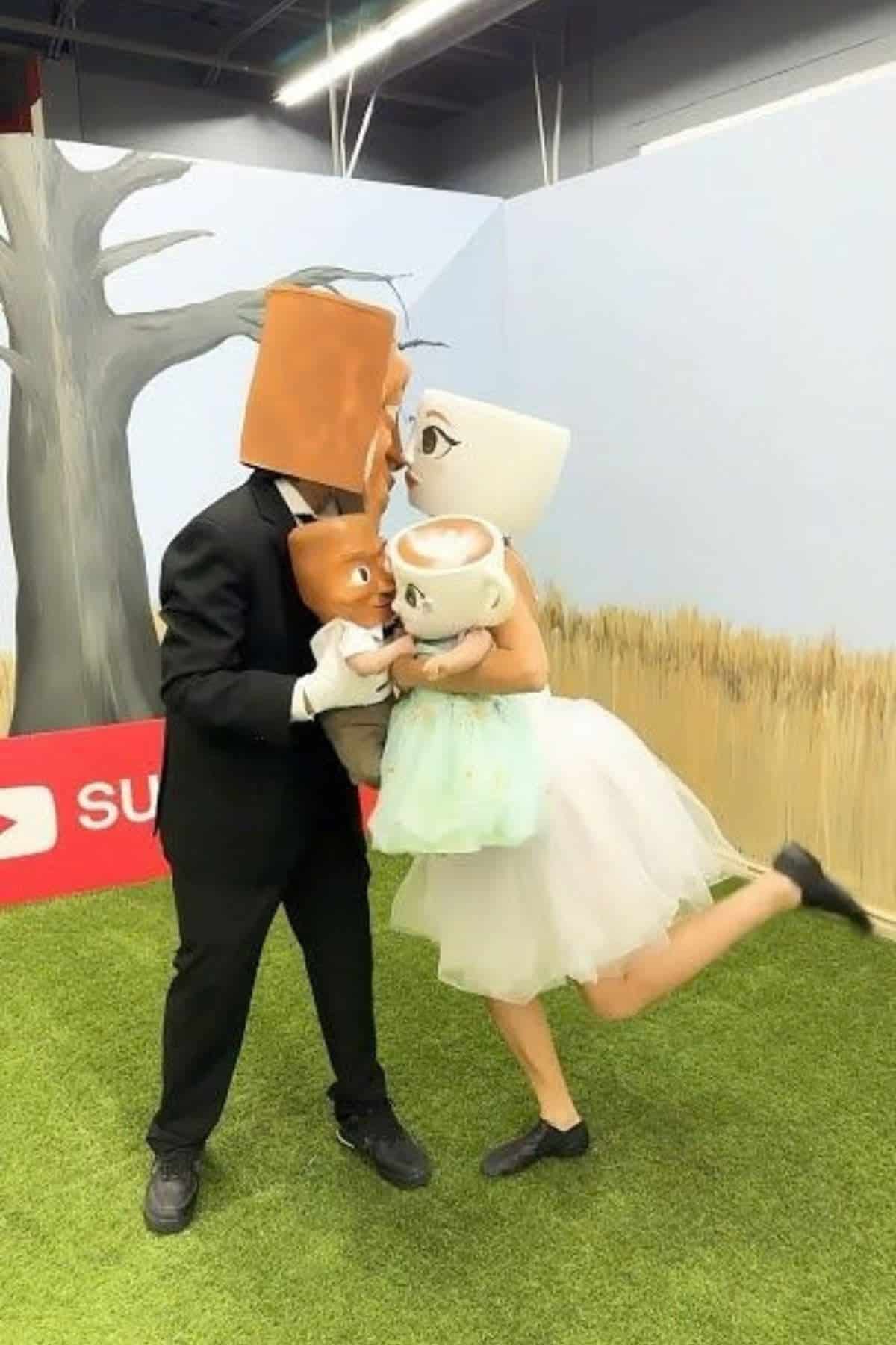 Two adults in cup and carton headpieces and formal wear hold two children with similar headpieces in a setting with artificial grass, a painted backdrop, and a red sign partly visible on the ground.