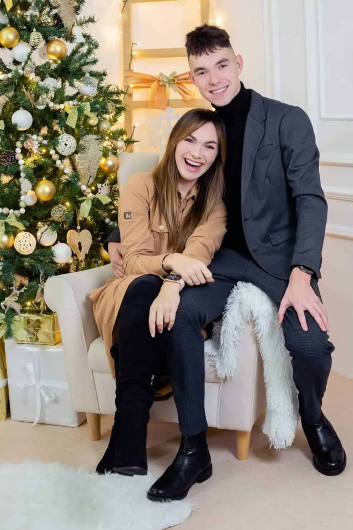 A young woman and man sit smiling together on a chair beside a decorated Christmas tree with gold and white ornaments and wrapped gifts.