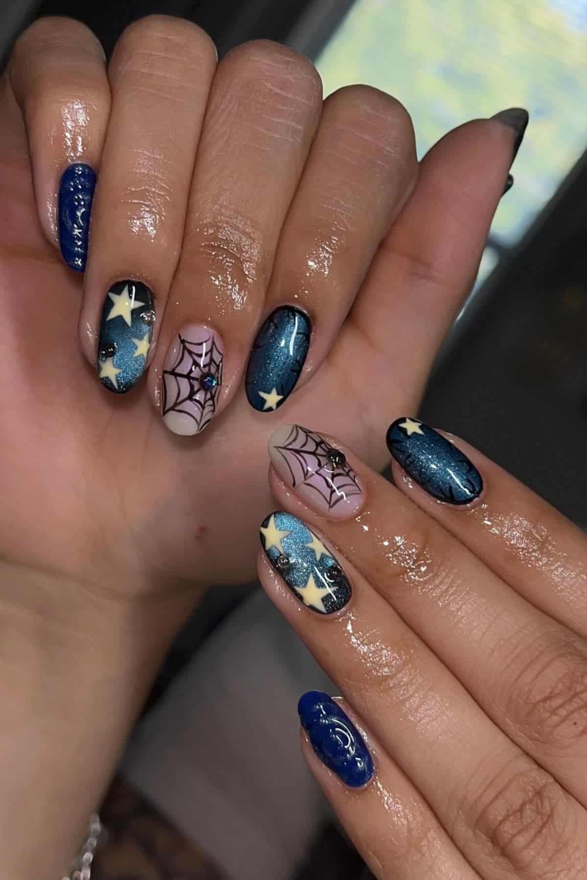 Hands with long, oval-shaped nails featuring blue polish, star designs, and spiderweb art. Nails appear shiny and freshly manicured.