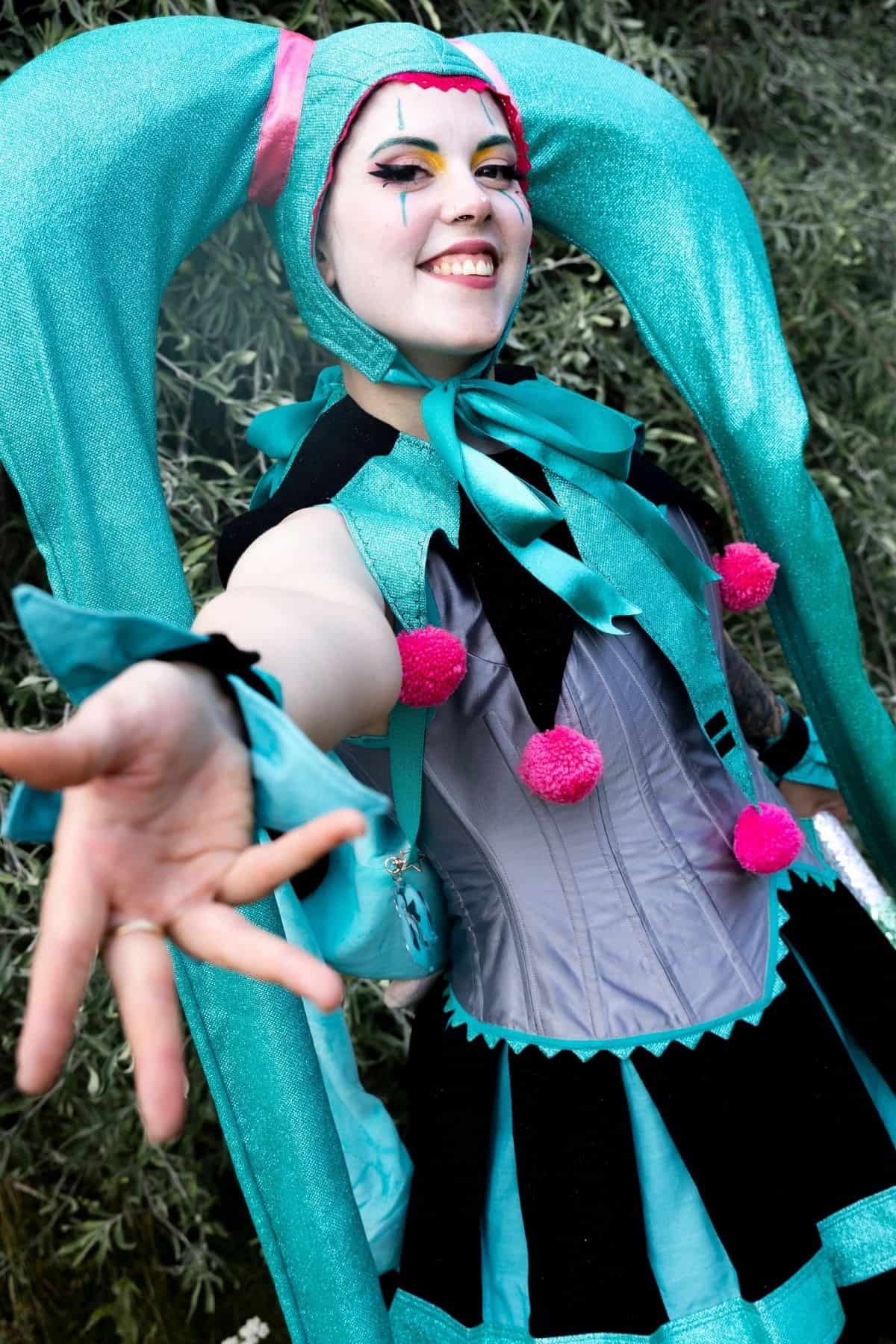 Person wearing a turquoise and black cosplay costume with large pigtails, pink pom-poms, and dramatic makeup, extending hand toward the camera, standing outdoors.