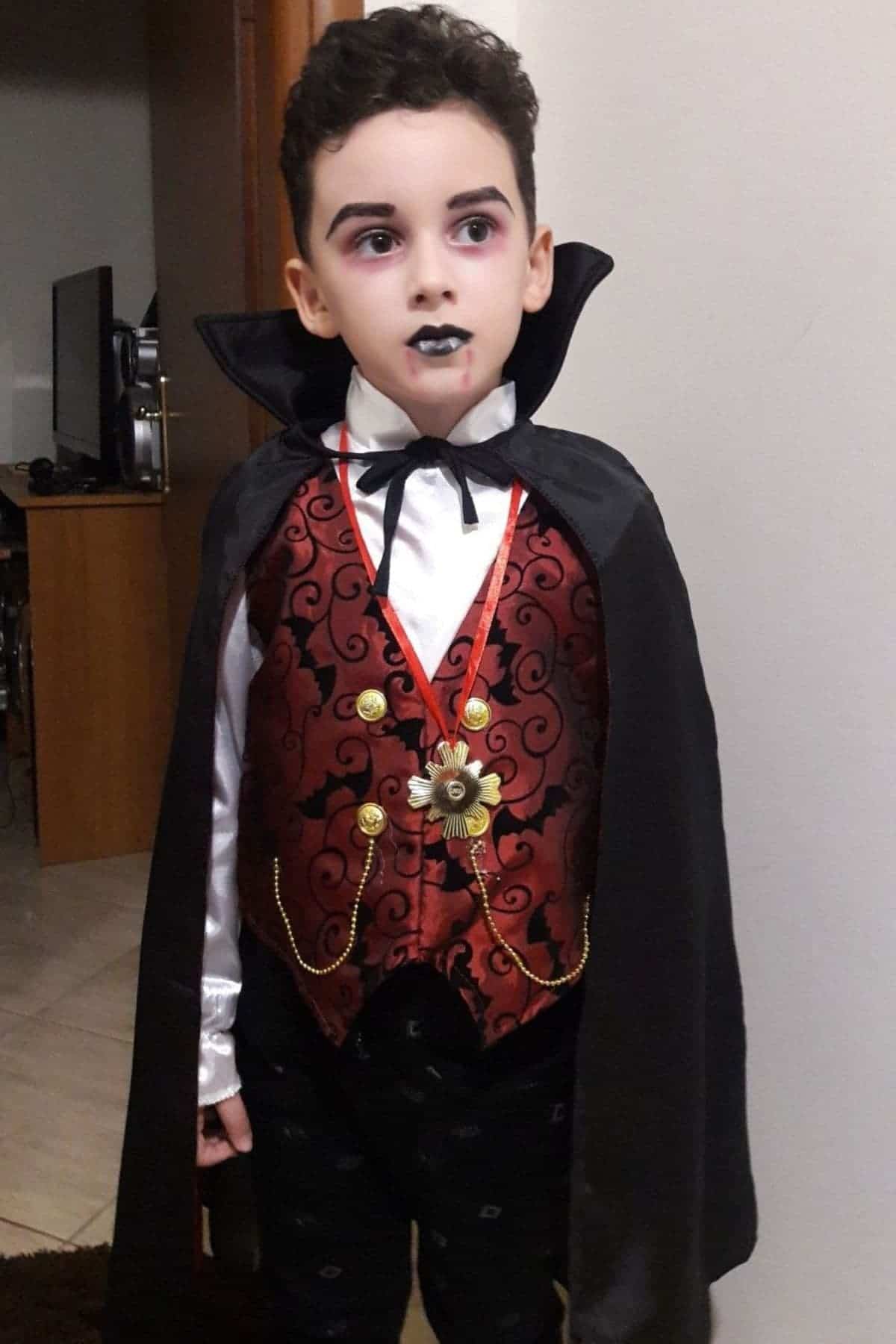 A child dressed as a vampire stands indoors, wearing a black cape, red vest with gold details, white shirt, and black lipstick.