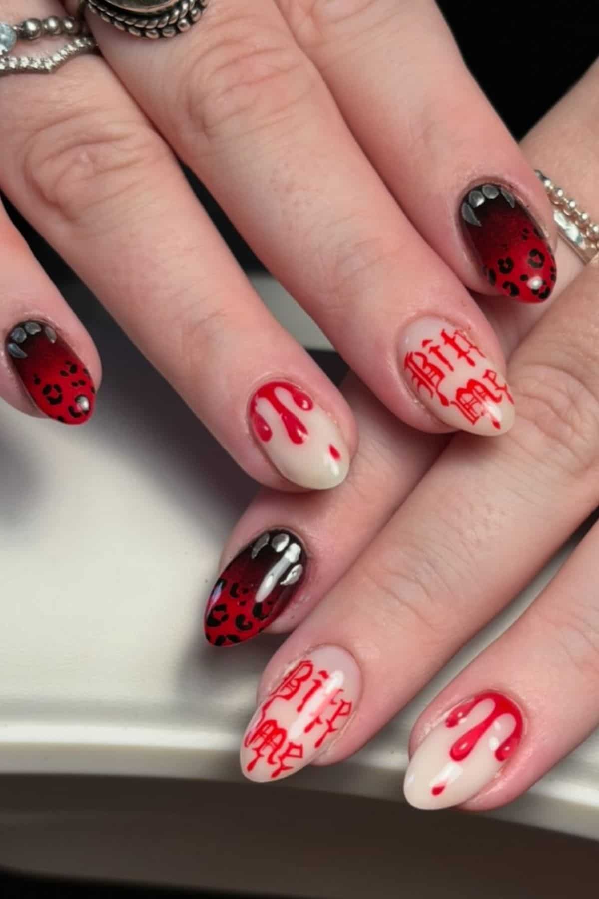 Close-up of hands with almond-shaped nails featuring red and black gradient, dripping blood designs, vampire fangs, and red writing resembling blood on a pale base.