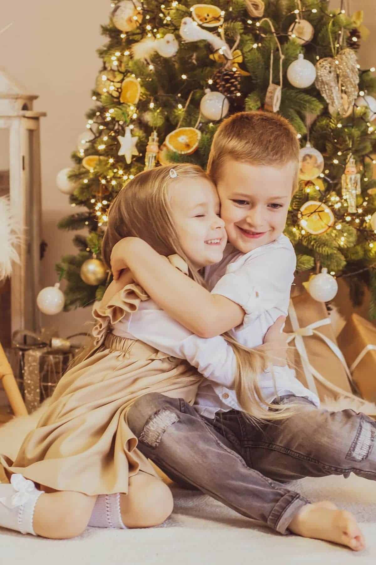 Two children, a boy and a girl, hug while sitting in front of a decorated Christmas tree with ornaments and lights.