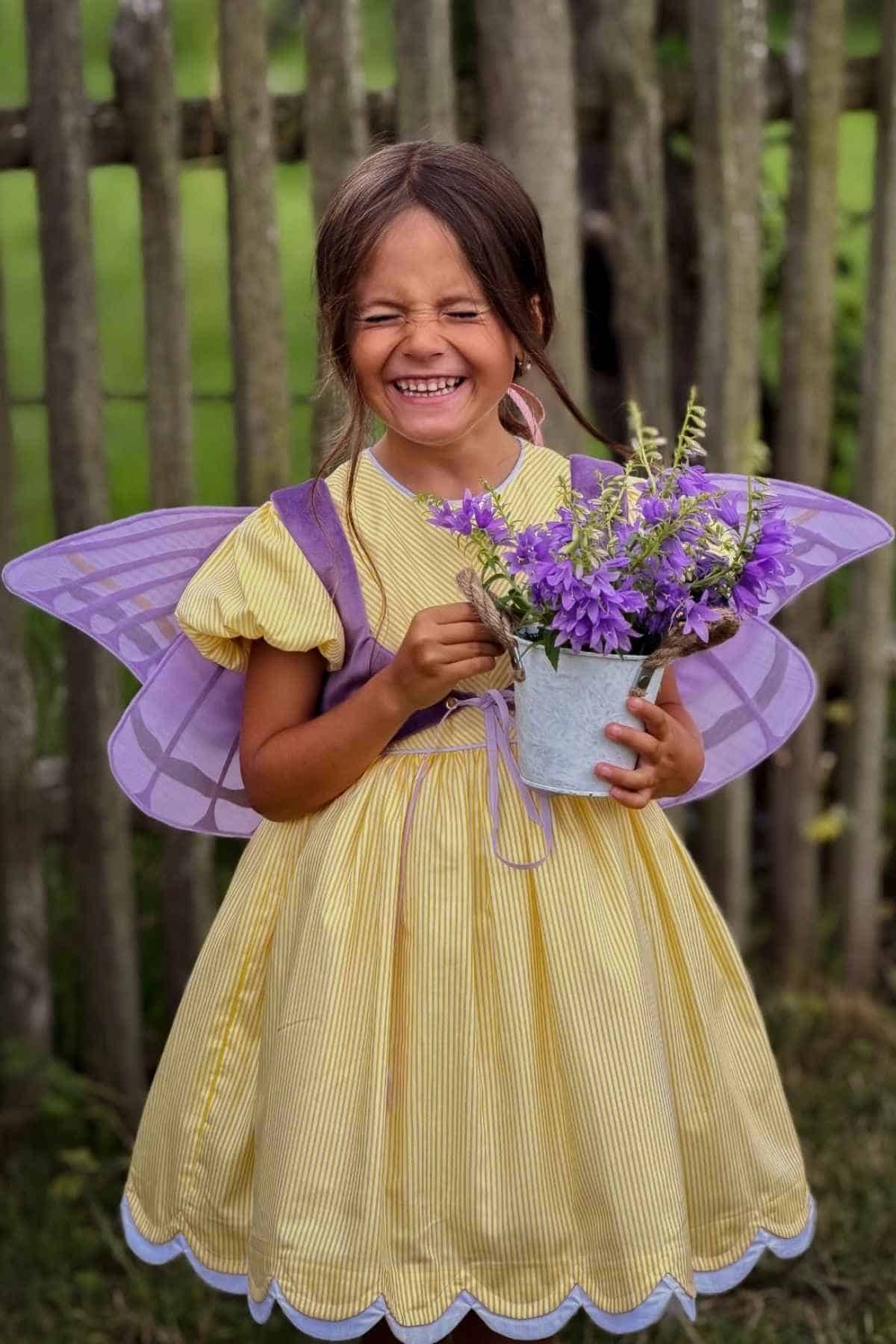 A smiling child in a yellow dress and purple fairy wings holds a metal bucket filled with purple flowers, standing in front of a wooden fence.