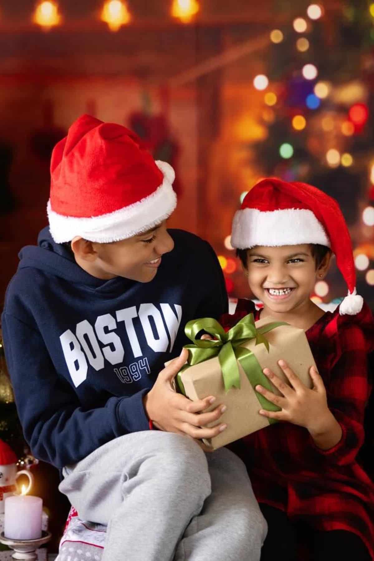 Two children wearing Santa hats sit together; the older child smiles at the younger, who is holding a wrapped gift with a green ribbon. Holiday decorations and lights are visible in the background.