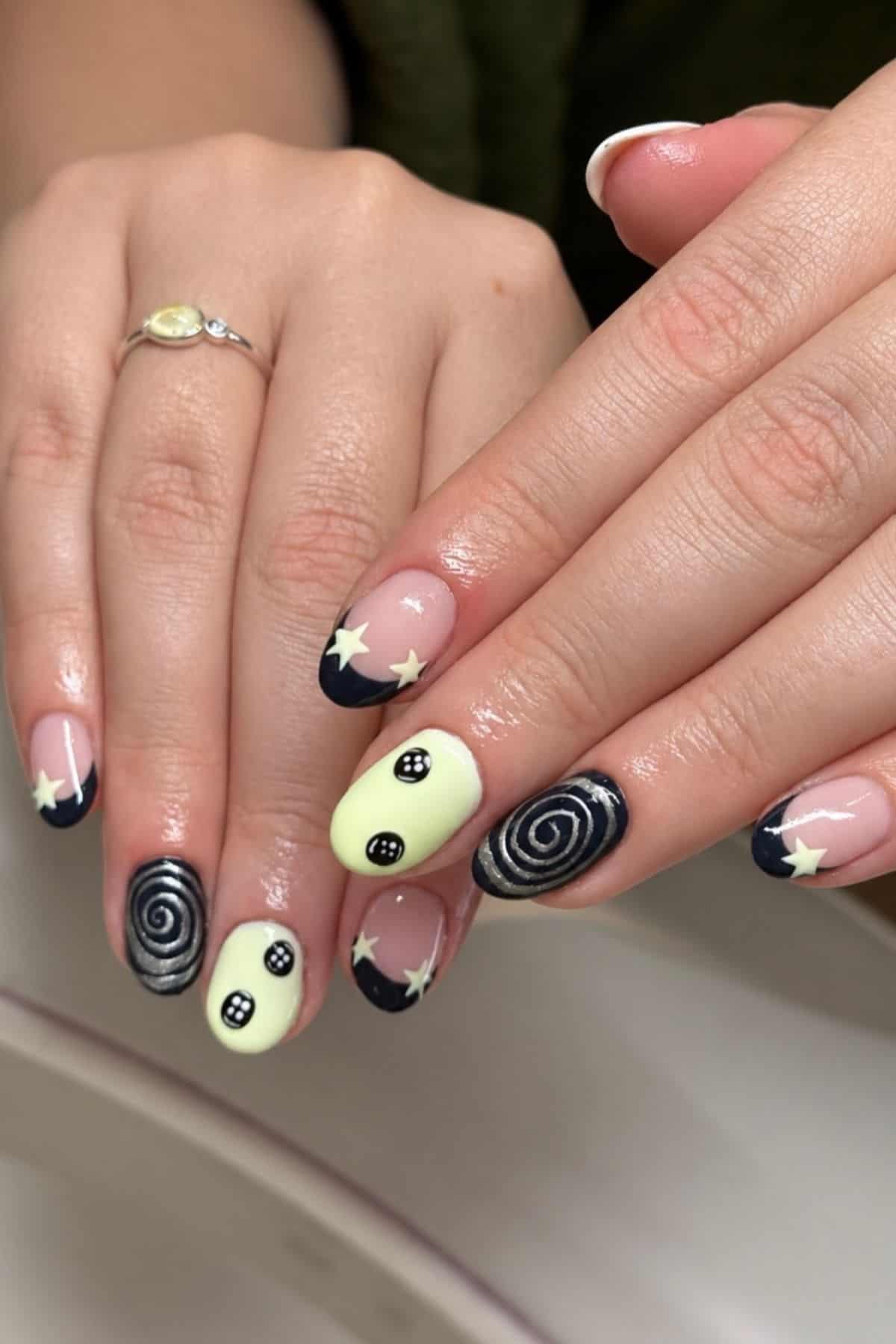 Close-up of hands with manicured nails featuring black swirls, yellow polish with button designs, and white star accents; person wears a silver ring with a light yellow stone.
