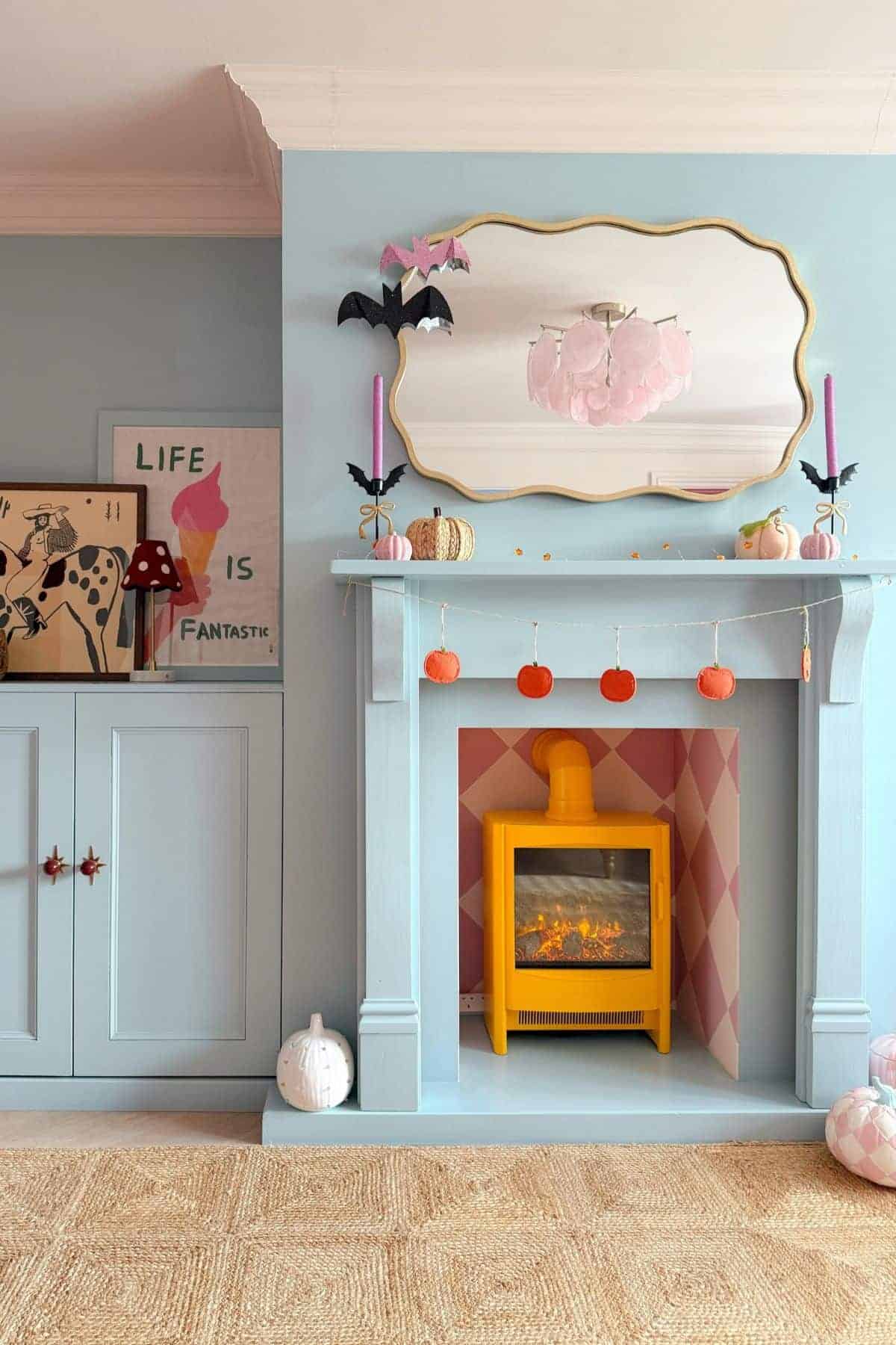 A pastel blue fireplace with a yellow electric heater, orange and white pumpkin decor, bat decorations, pink candles, and a gold-framed mirror above.