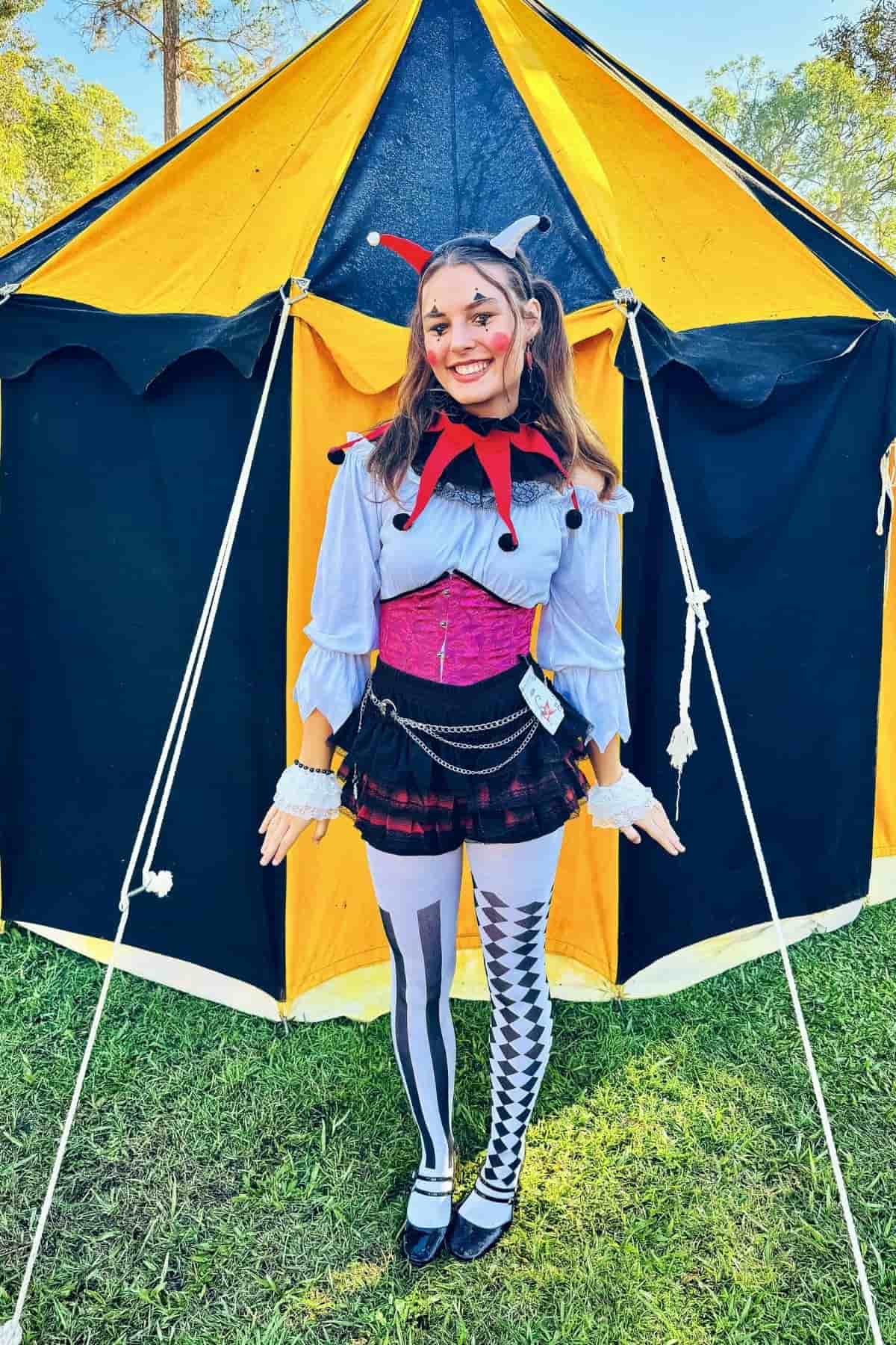 Person in a jester costume stands smiling in front of a yellow and black tent on grass, wearing a red collar, corset, and mismatched tights.