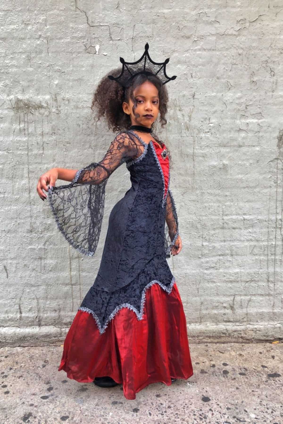 Child in a red and black costume dress with lace sleeves and a black crown headpiece poses against a textured gray wall.