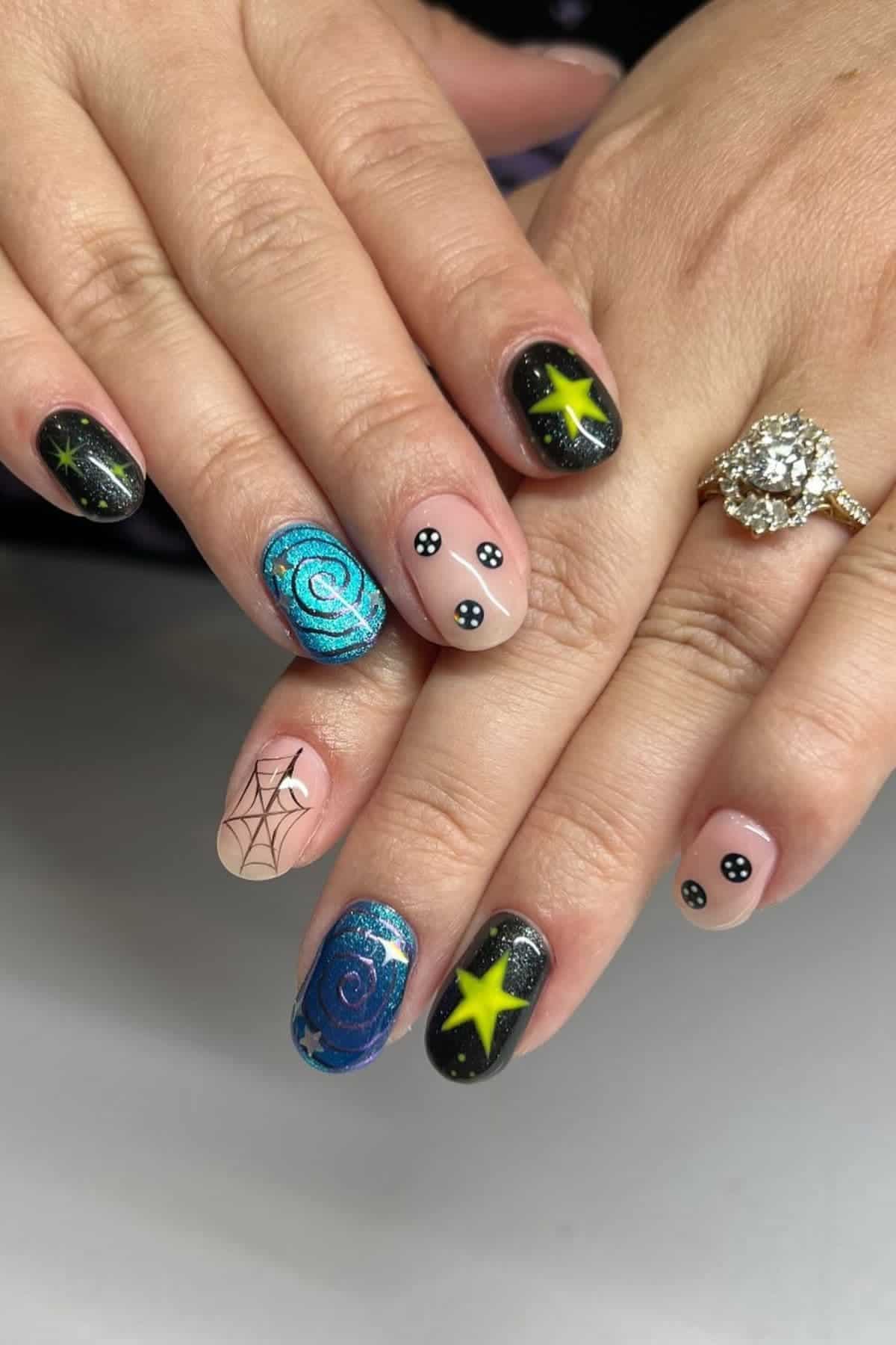Hands with manicured nails featuring galaxy designs, stars, swirls, spiderweb, and black nails with white dots. Person wears a ring with a large circular gem.