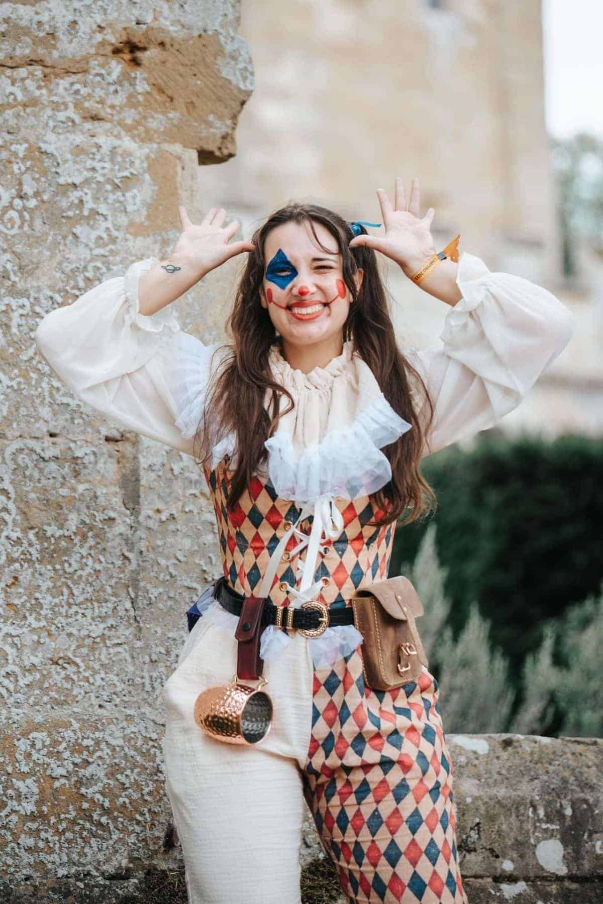Person in a harlequin costume with colorful face paint stands outdoors, smiling and holding their hands by their head with fingers spread.