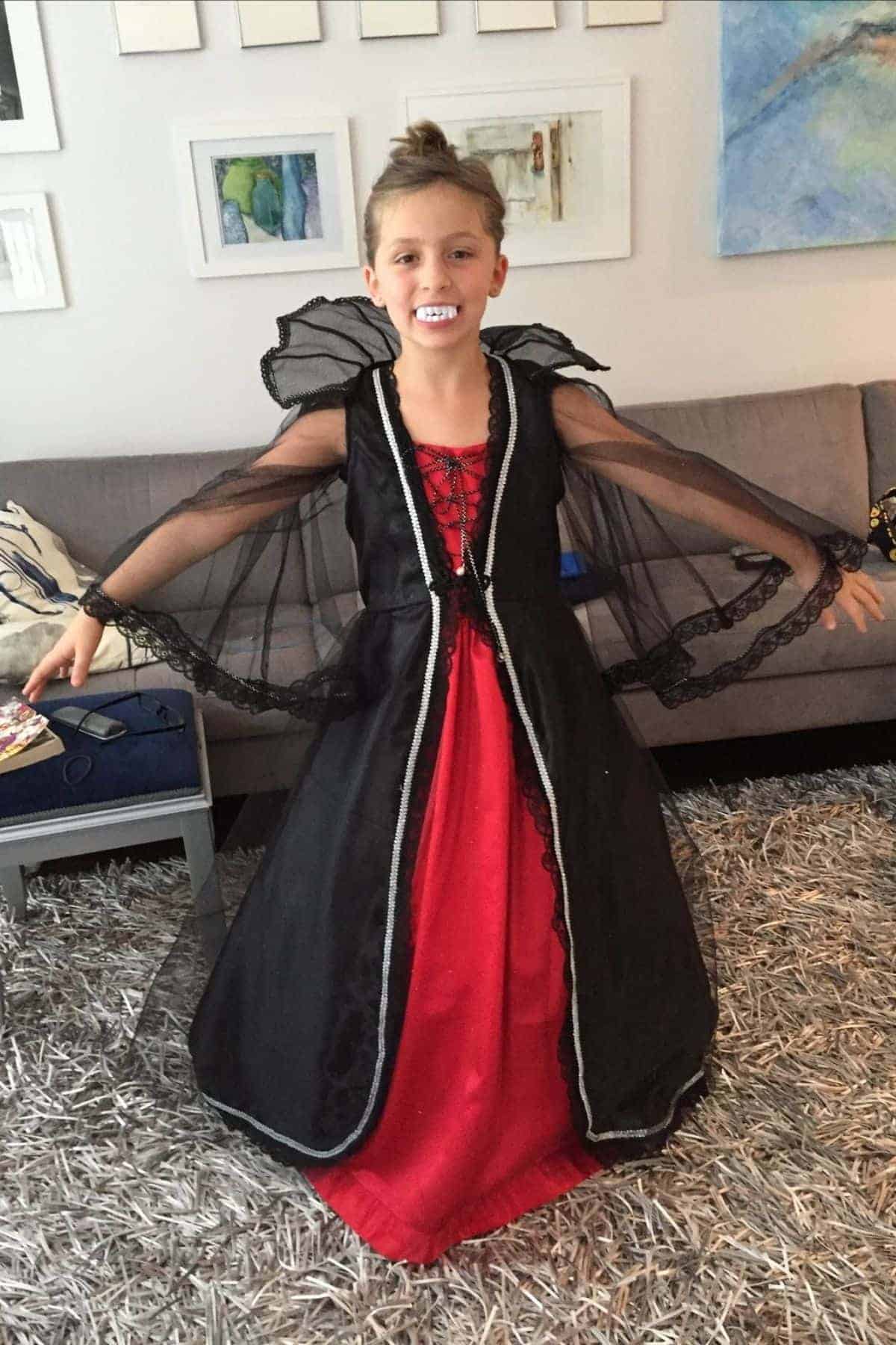 A child stands indoors wearing a red and black vampire costume dress with mesh sleeves, smiling with arms outstretched.