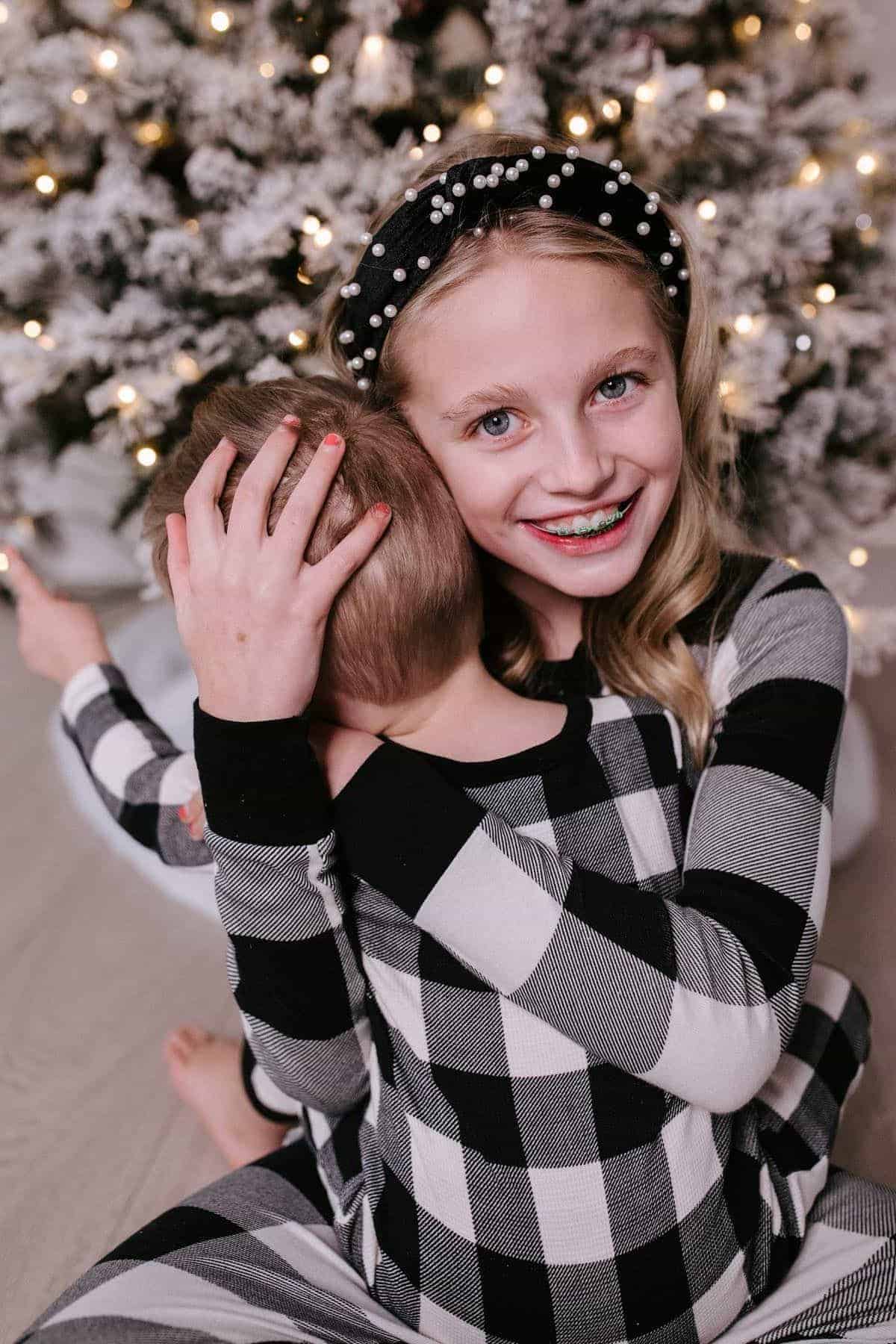 Two children in matching plaid pajamas sit in front of a lit Christmas tree. The older child smiles at the camera while hugging the younger child, who faces away.