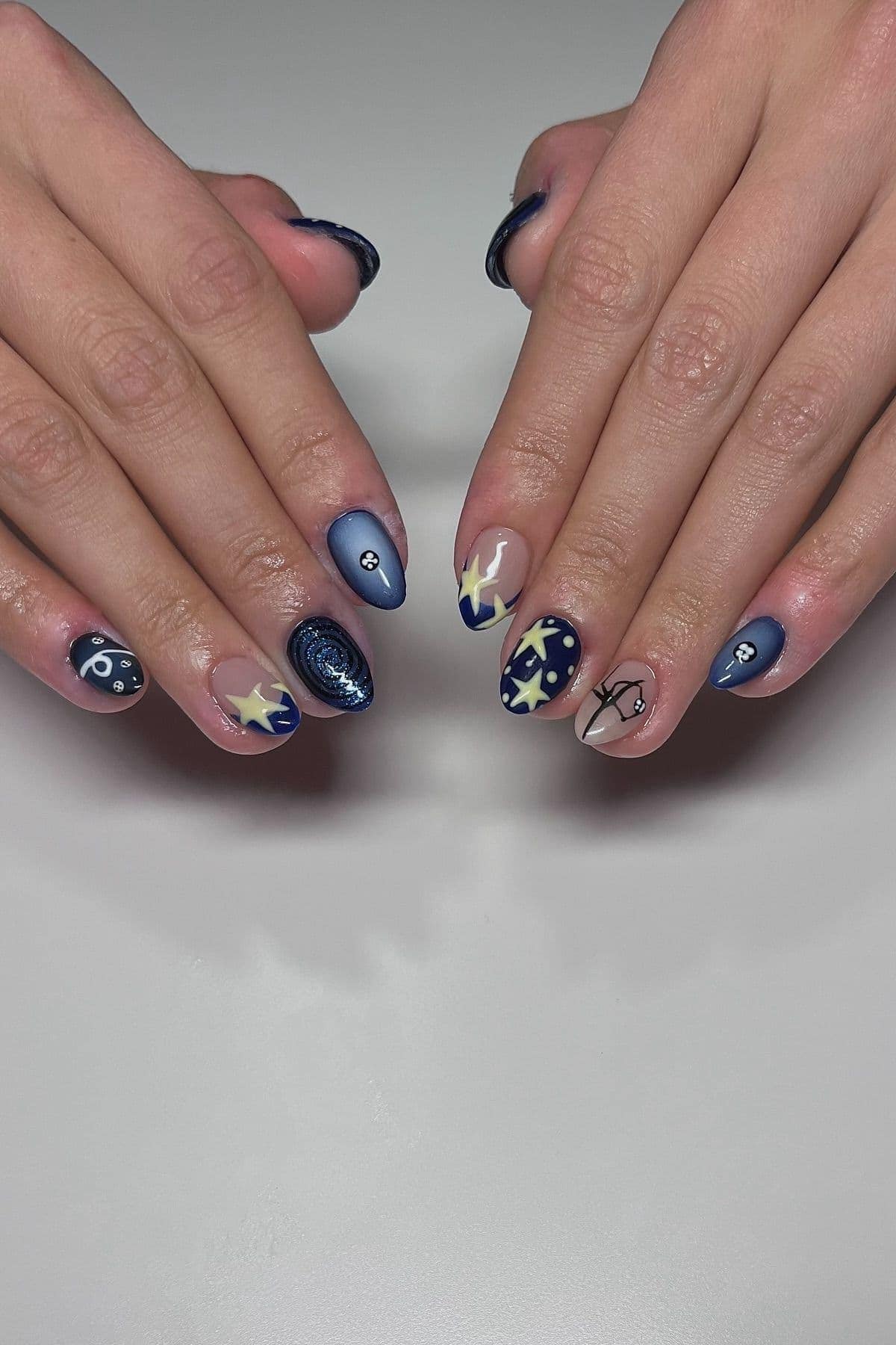 Two hands with navy blue, star, moon, and zodiac-themed nail art designs on almond-shaped nails, displayed on a light grey surface.