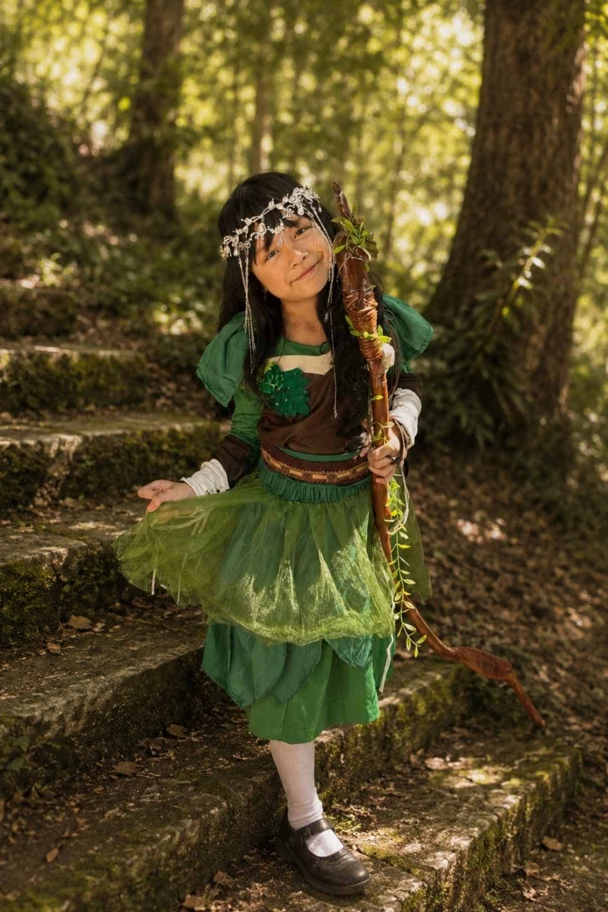 A young child in a green costume with a flower crown and a wooden staff stands on mossy stone steps in a forested area.