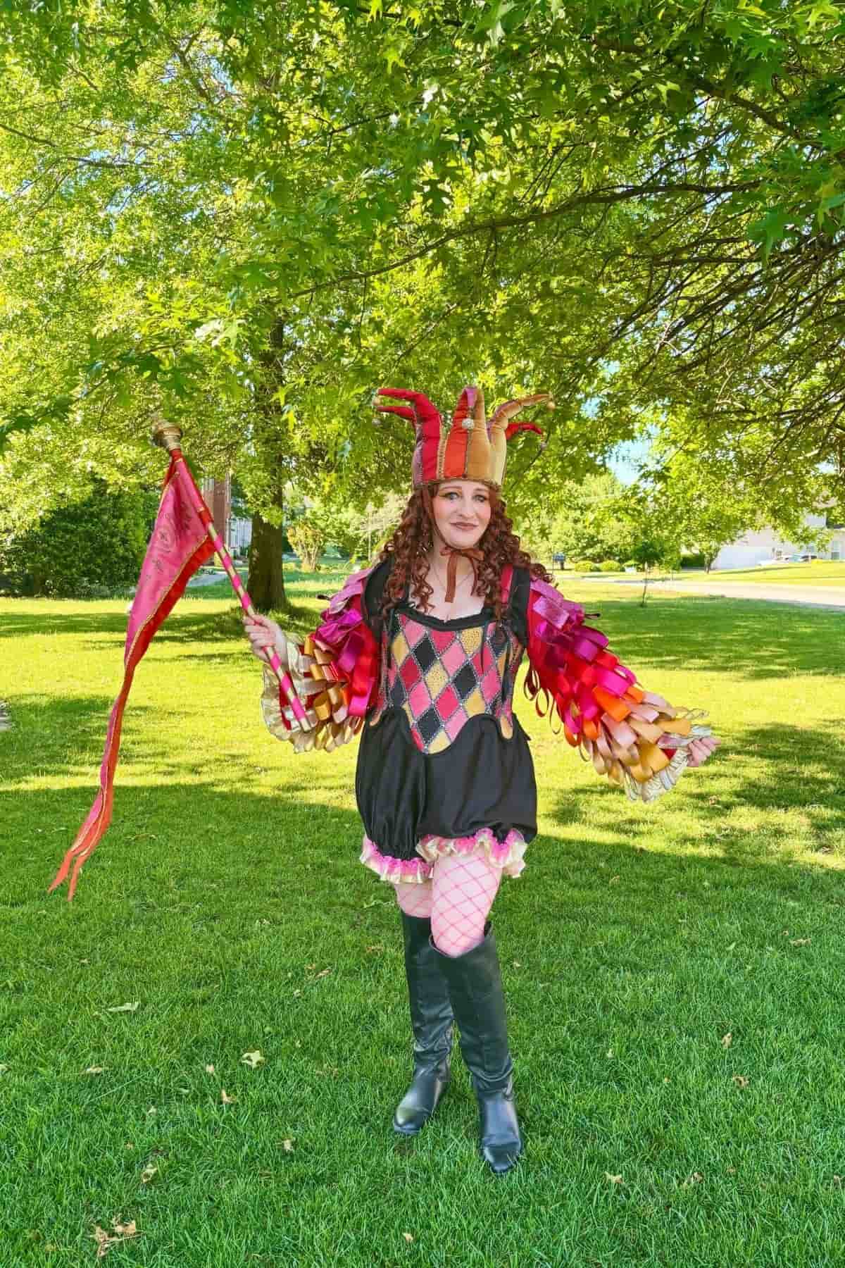 Person in a colorful jester costume with a red flag stands on green grass under leafy trees on a sunny day.