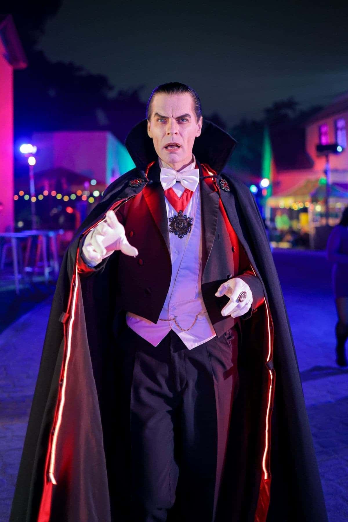 A person dressed as a classic vampire with a black and red cape, white gloves, and formal attire stands outdoors at night with colorful lights in the background.