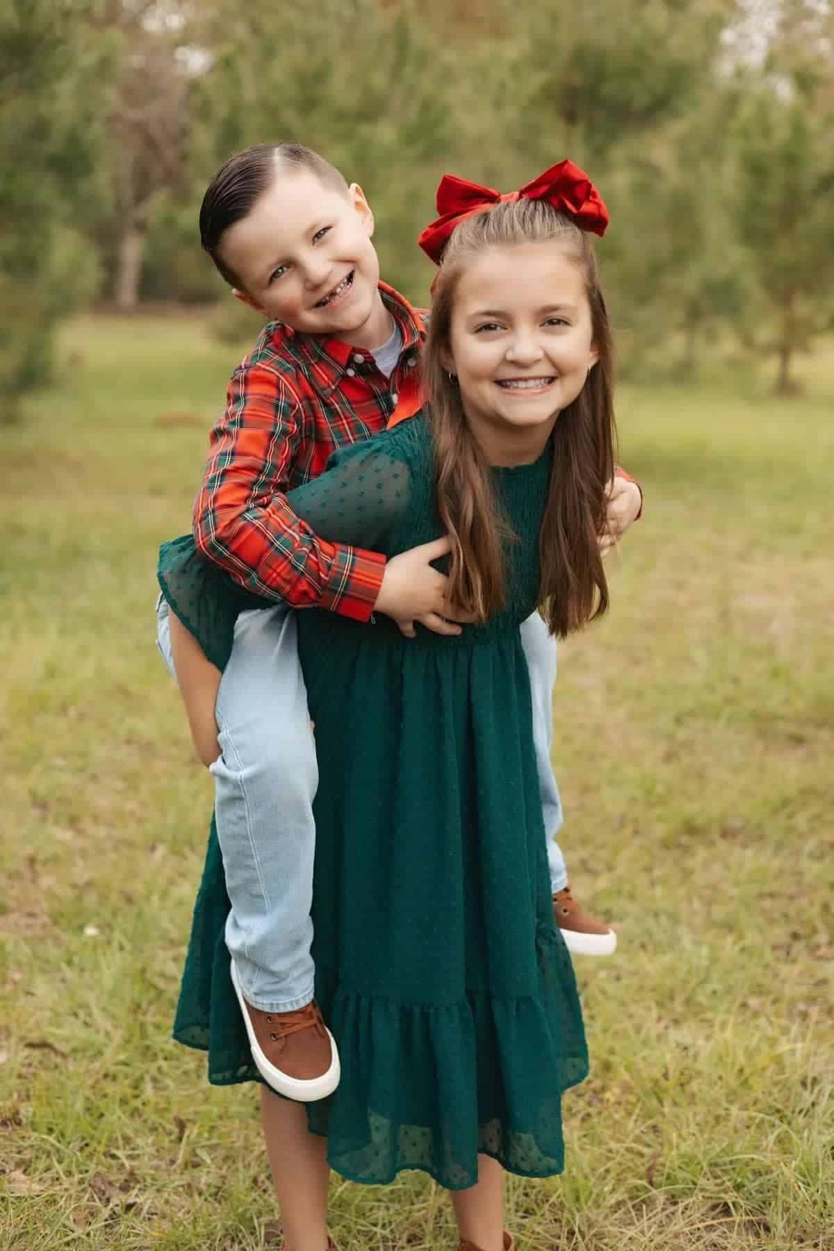 A girl in a green dress gives a piggyback ride to a boy in a red plaid shirt as they smile outdoors on grass with trees in the background.
