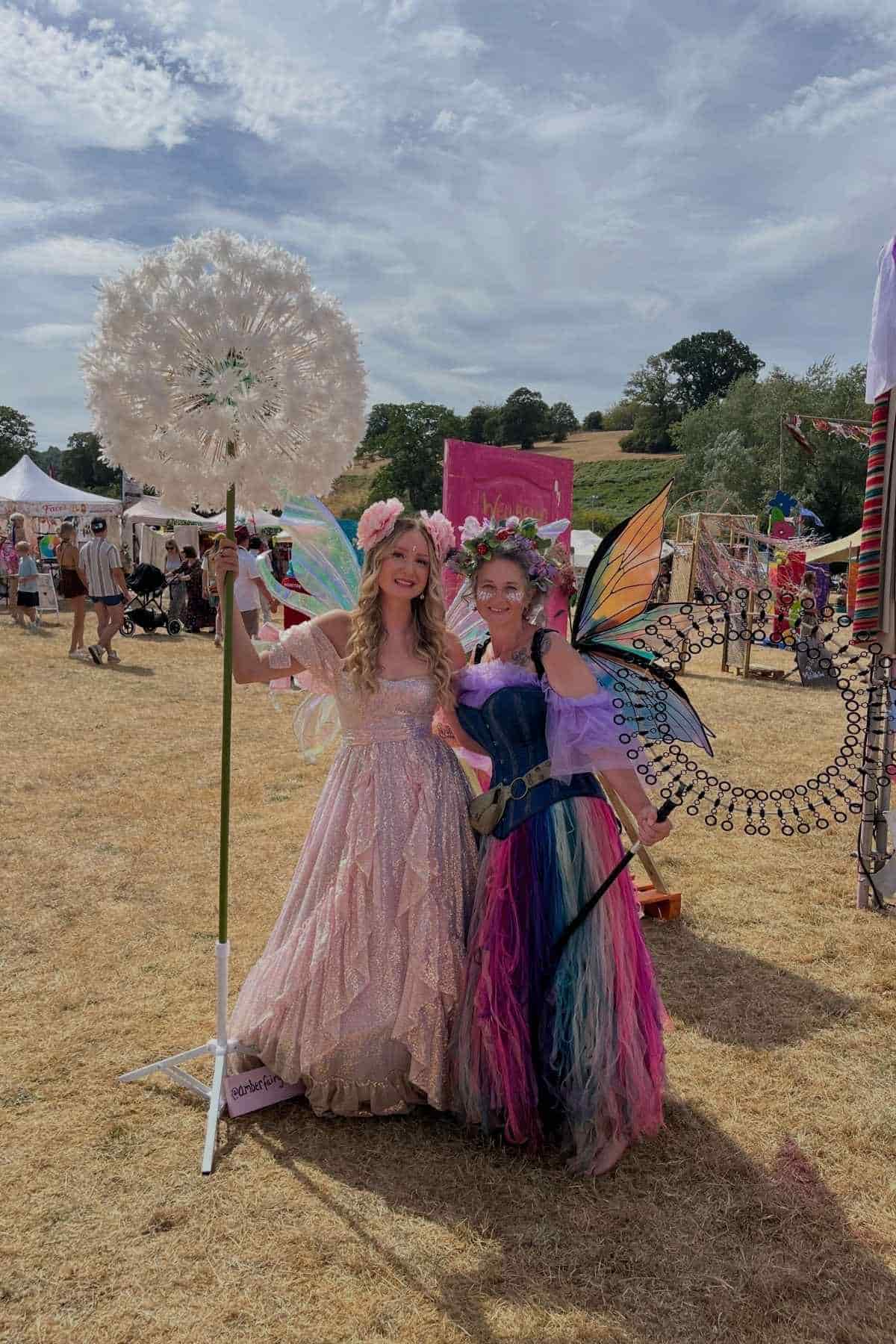 Two people dressed in elaborate fairy costumes stand outdoors at a festival, one holding a large artificial dandelion, with tents and festivalgoers visible in the background.