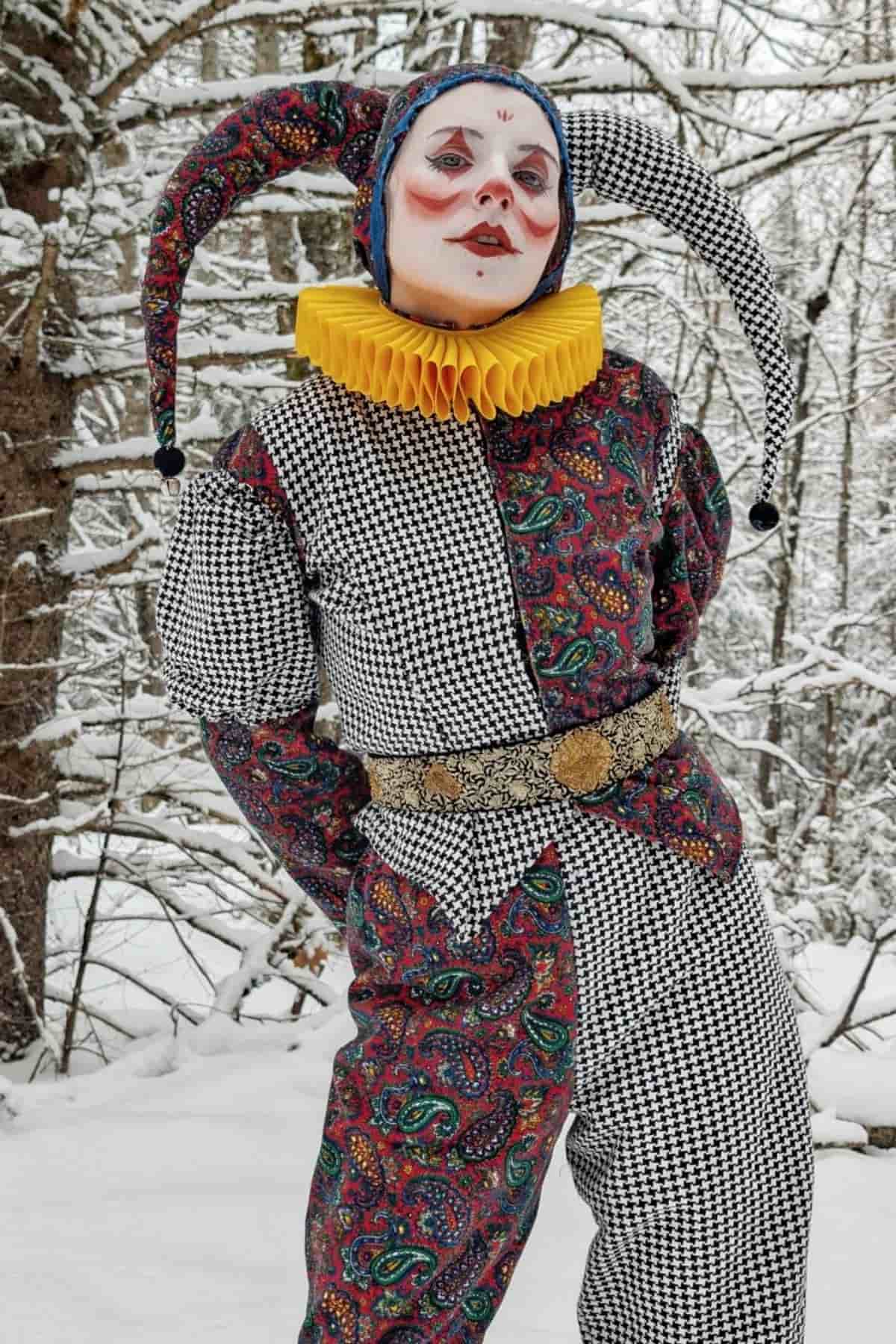 Person in a jester costume with a ruffled yellow collar stands in a snowy forest. The costume features black-and-white checks and paisley patterns, with face paint and a pointed hat.