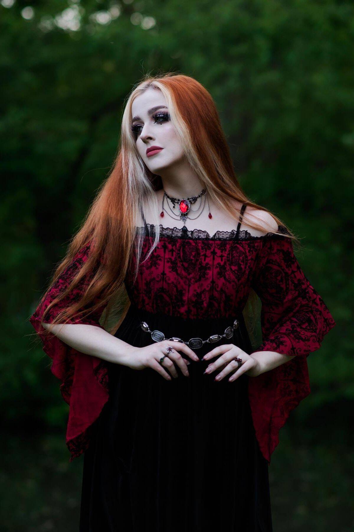 A woman with long red hair wears a burgundy and black gothic dress and jewelry, standing outdoors against a blurred green background.