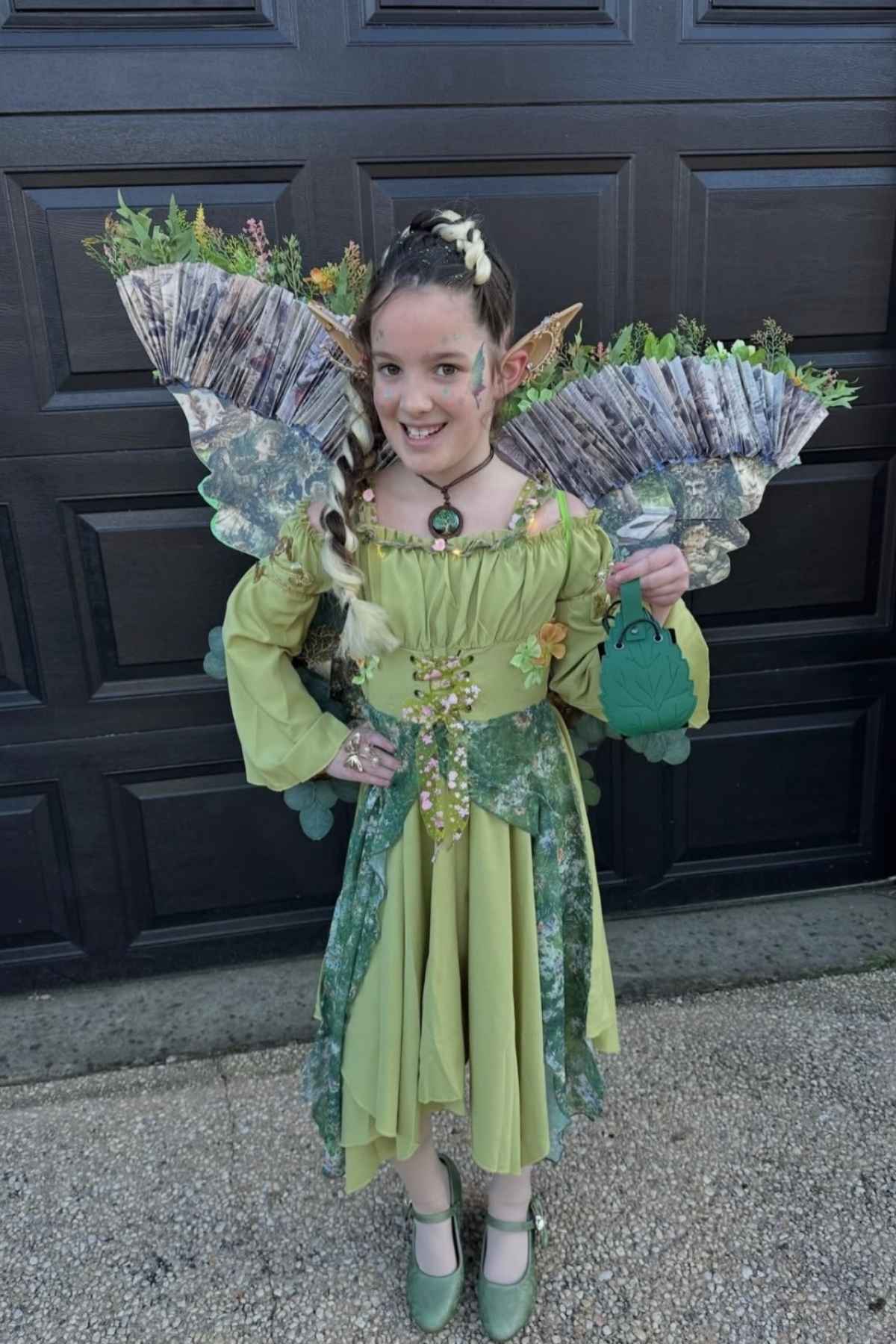A child in a green fairy costume with large decorative wings stands outdoors in front of a dark garage door, holding a green bag and smiling at the camera.